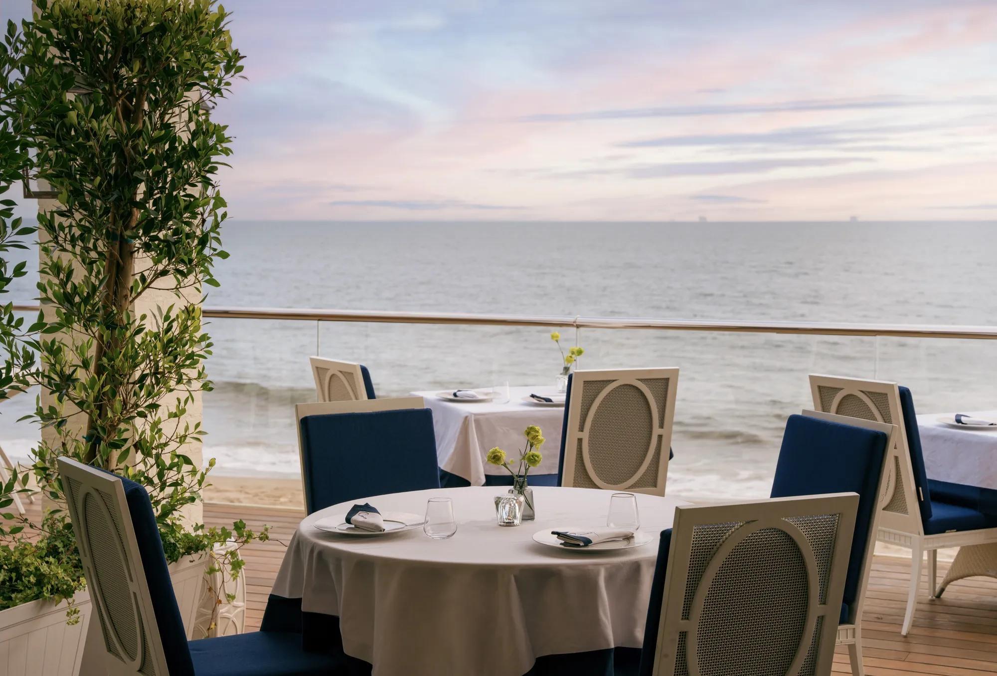 An outdoor restaurant terrace with round tables set for dinner, overlooking the ocean at dusk.