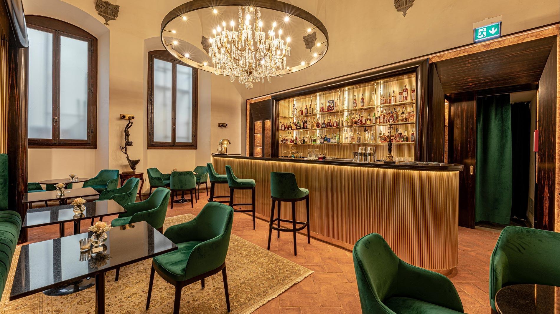 a hotel bar with a backlit gold shelf of bottles, green velvet chairs and stools, and a round crystal chandelier