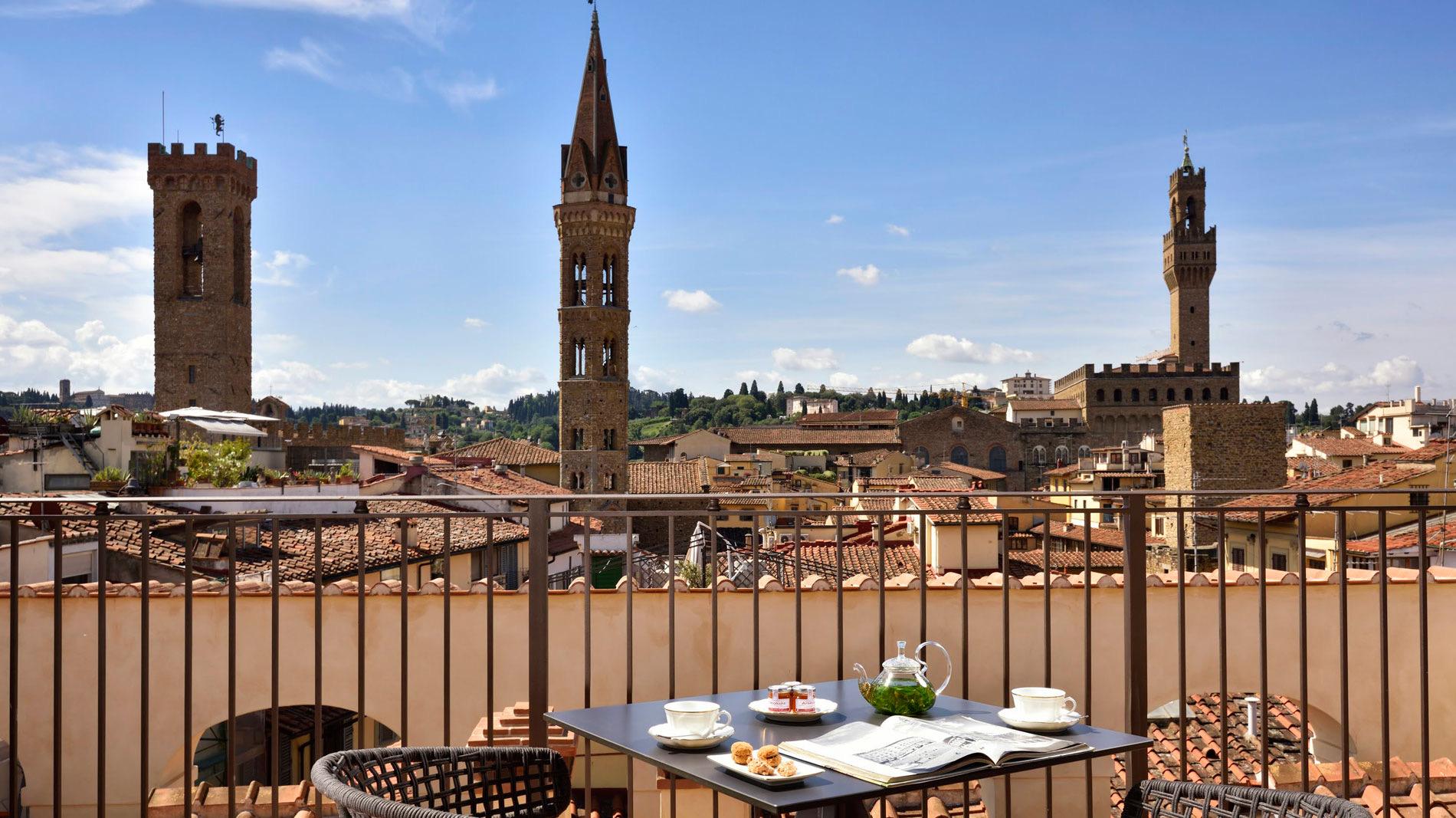 a rooftop terrace table set with tea and pastries, a wrought iron railing in front, and three medieval Florence towers against a blue sky