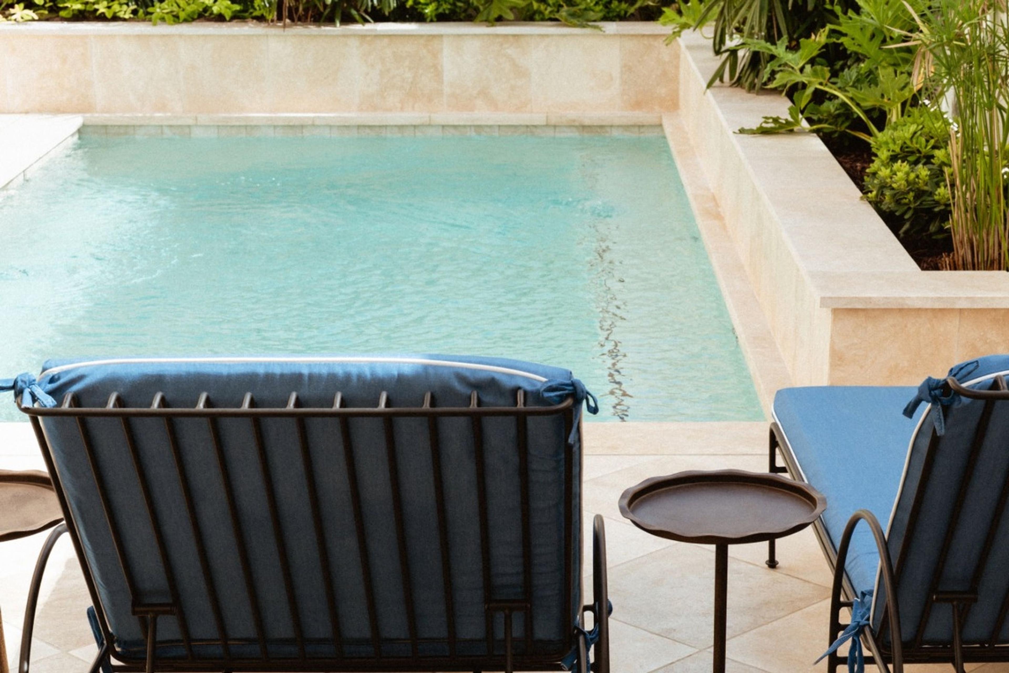 Two navy cushioned lounge chairs and a small side table beside a tiled plunge pool, with planting along the back wall.