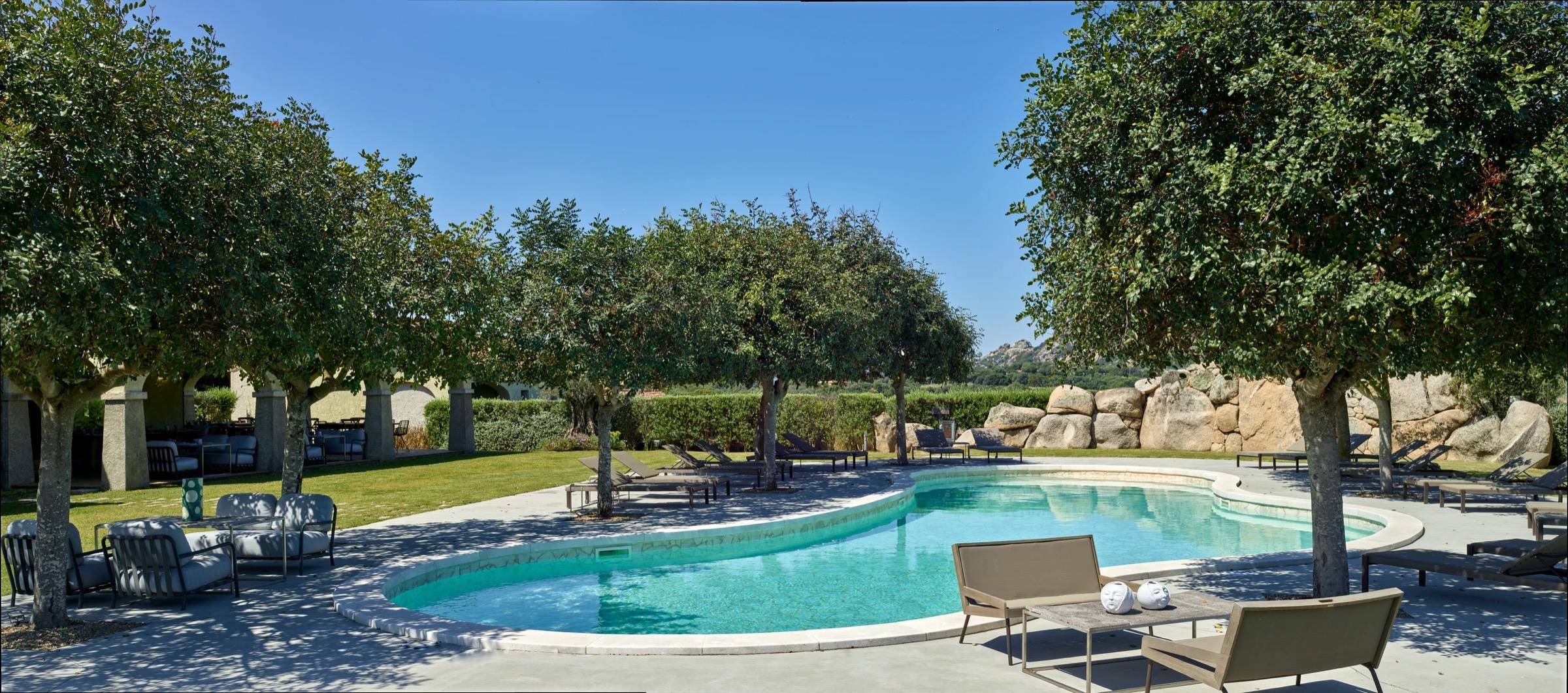 a curved swimming pool surrounded by sun loungers and shade trees, with boulders and a lawn beyond