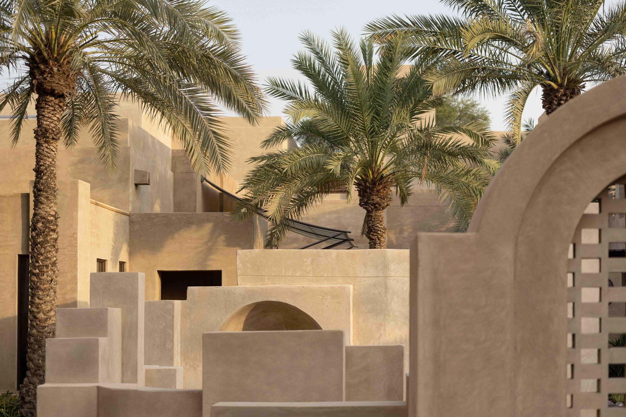 Angular plastered walls and an arched gateway rise among date palms, lit by late afternoon sun.