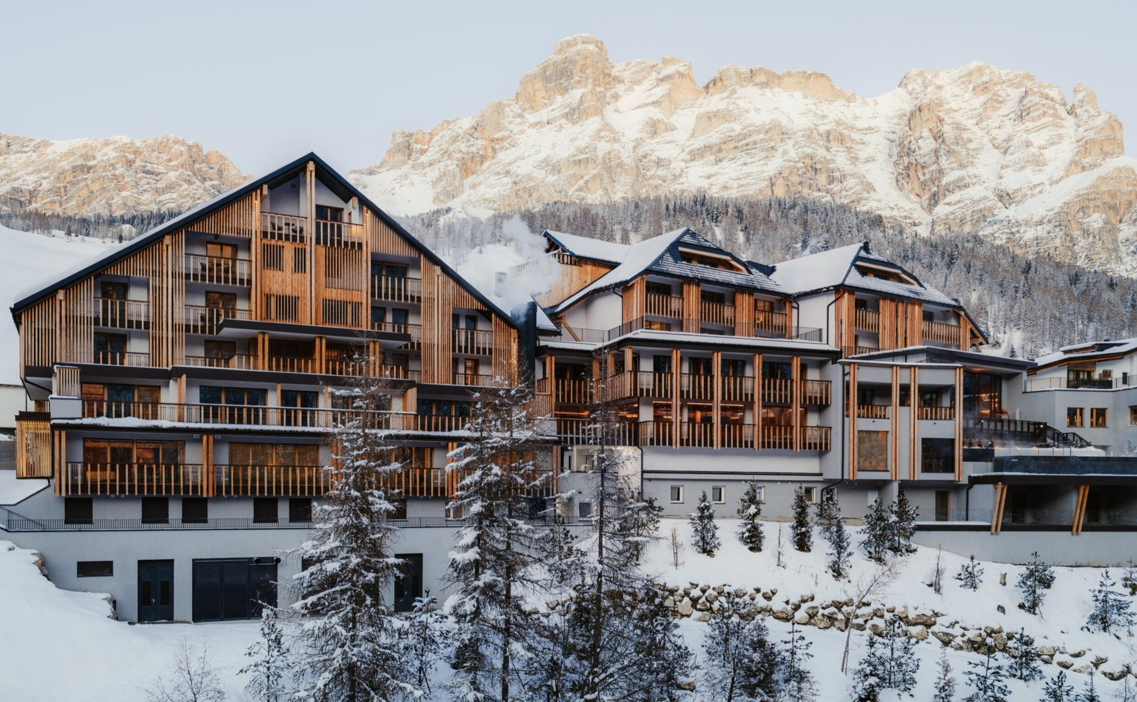 a timber and stone hotel building with snow covered balconies, under jagged mountain peaks lit gold by afternoon sun
