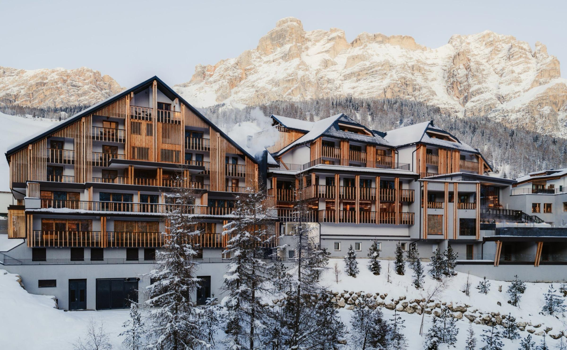 a timber and stone hotel building with snow covered balconies, under jagged mountain peaks lit gold by afternoon sun
