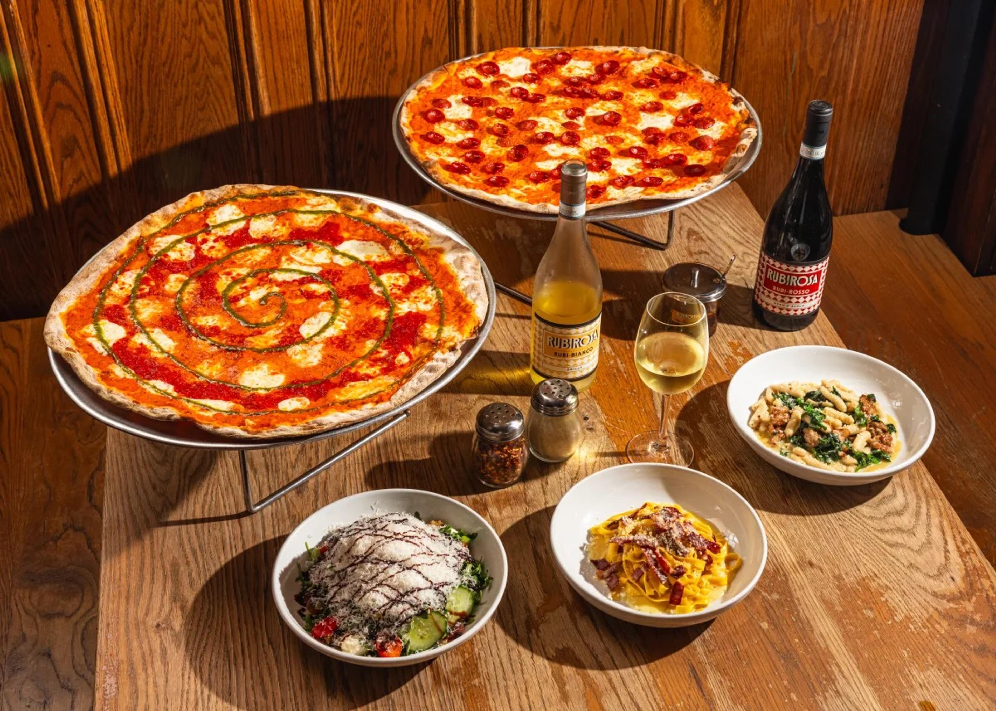 A thin-crust pizza on a raised stand among plates of pasta and salad and a bottle of wine