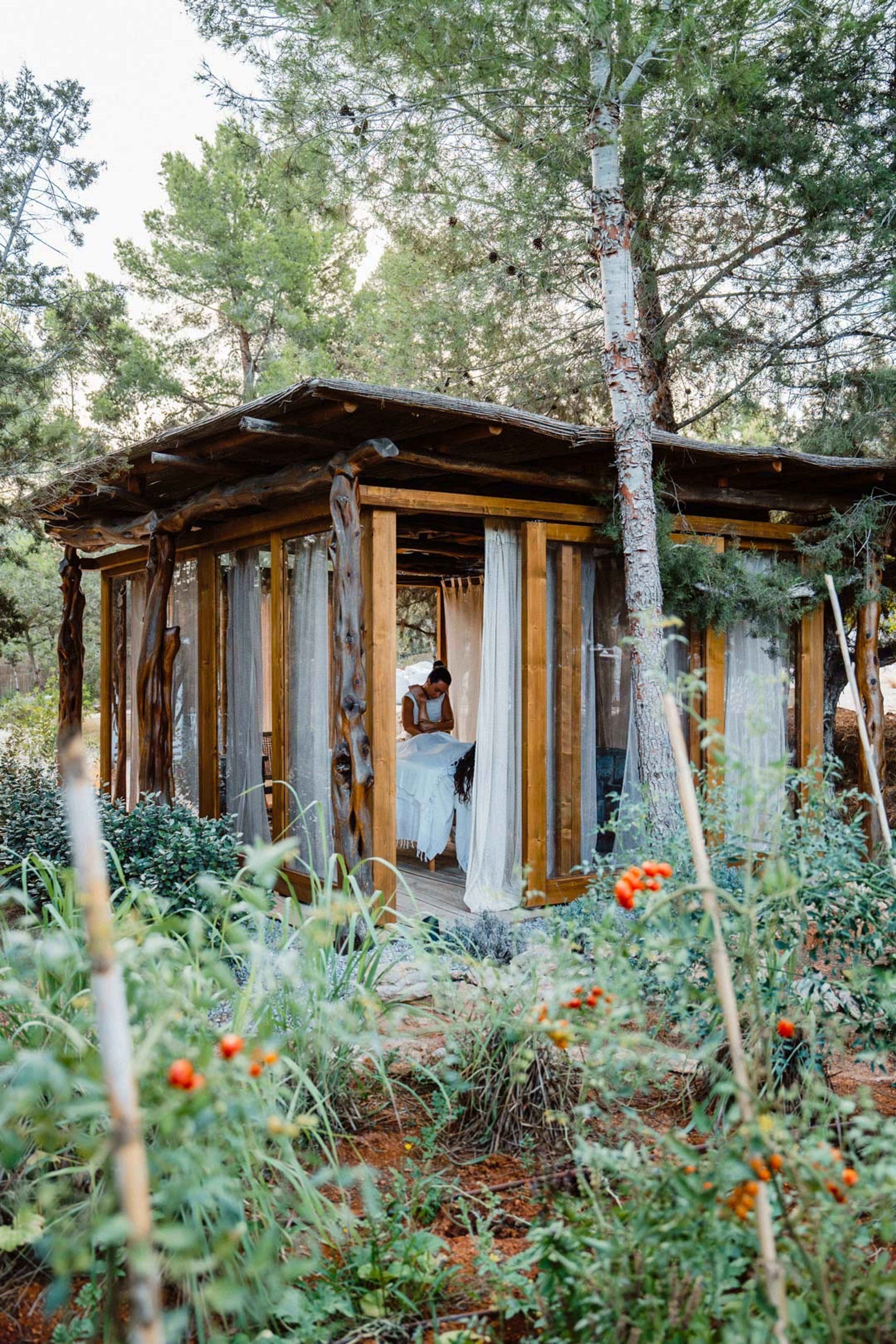 An open-sided wooden pavilion with sheer curtains, built among pine trees, with a person preparing a massage table inside.