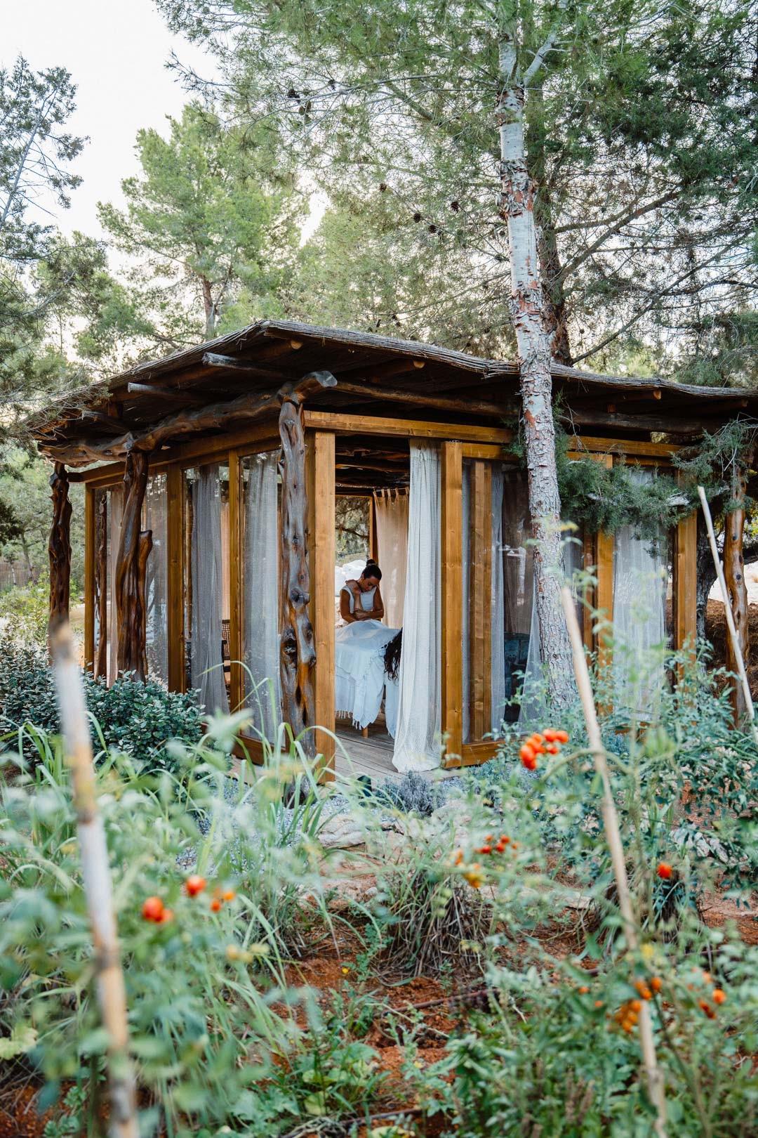 An open-sided wooden pavilion with sheer curtains, built among pine trees, with a person preparing a massage table inside.