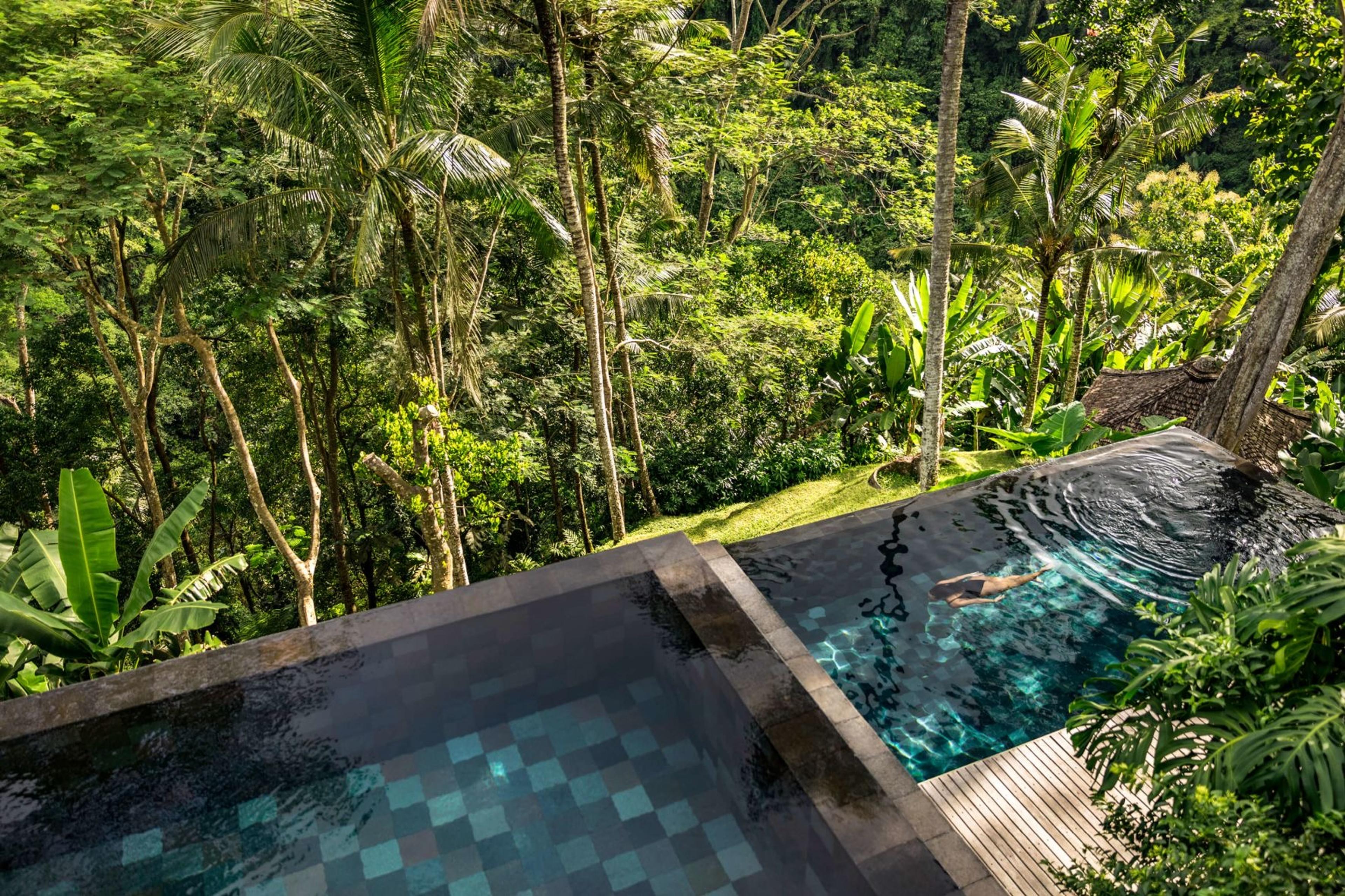 An aerial view looking down into two adjoining tiled pools set against a jungle backdrop, with a person swimming underwater.