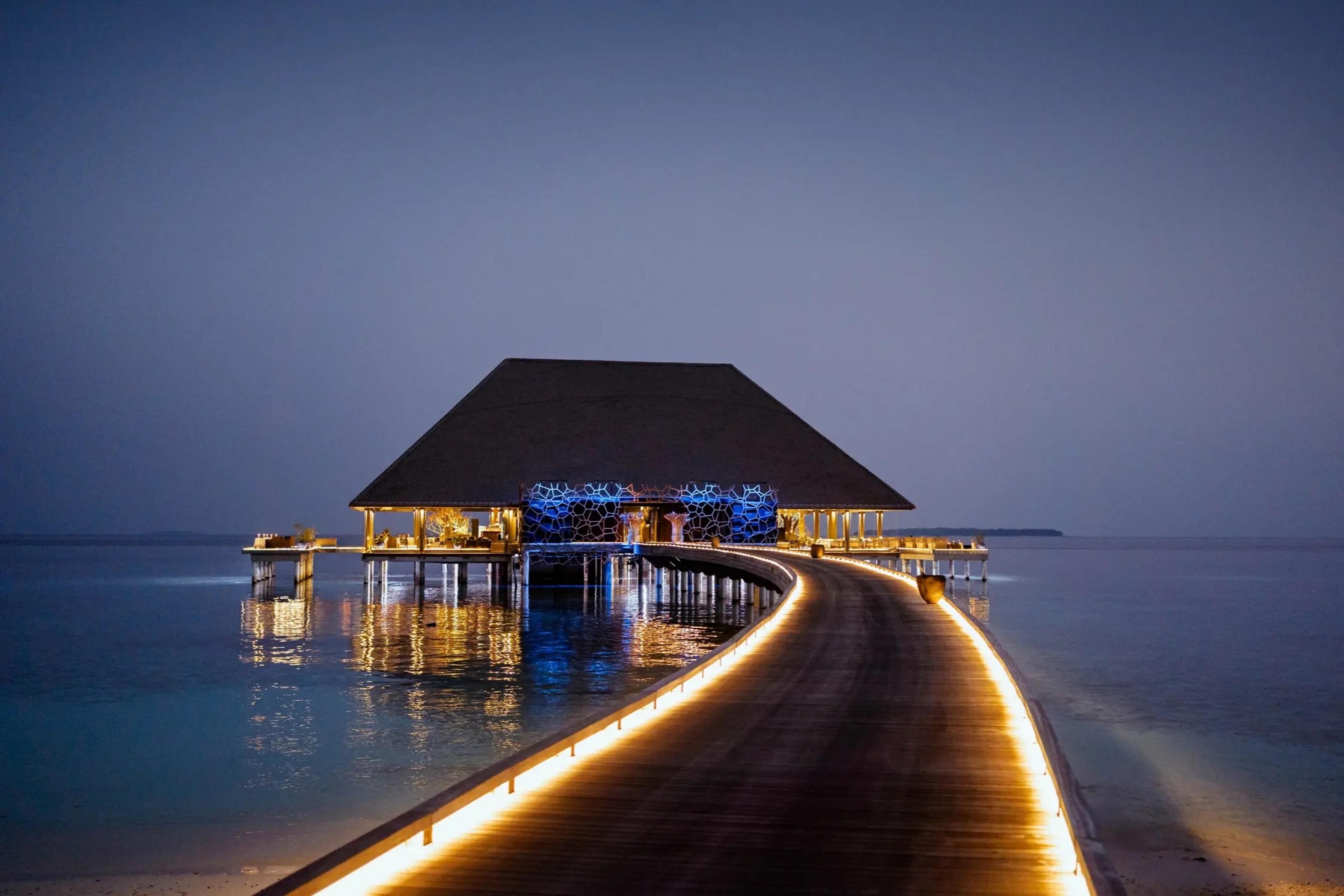 An overwater restaurant lit at dusk, reached by a long lit walkway across the lagoon.