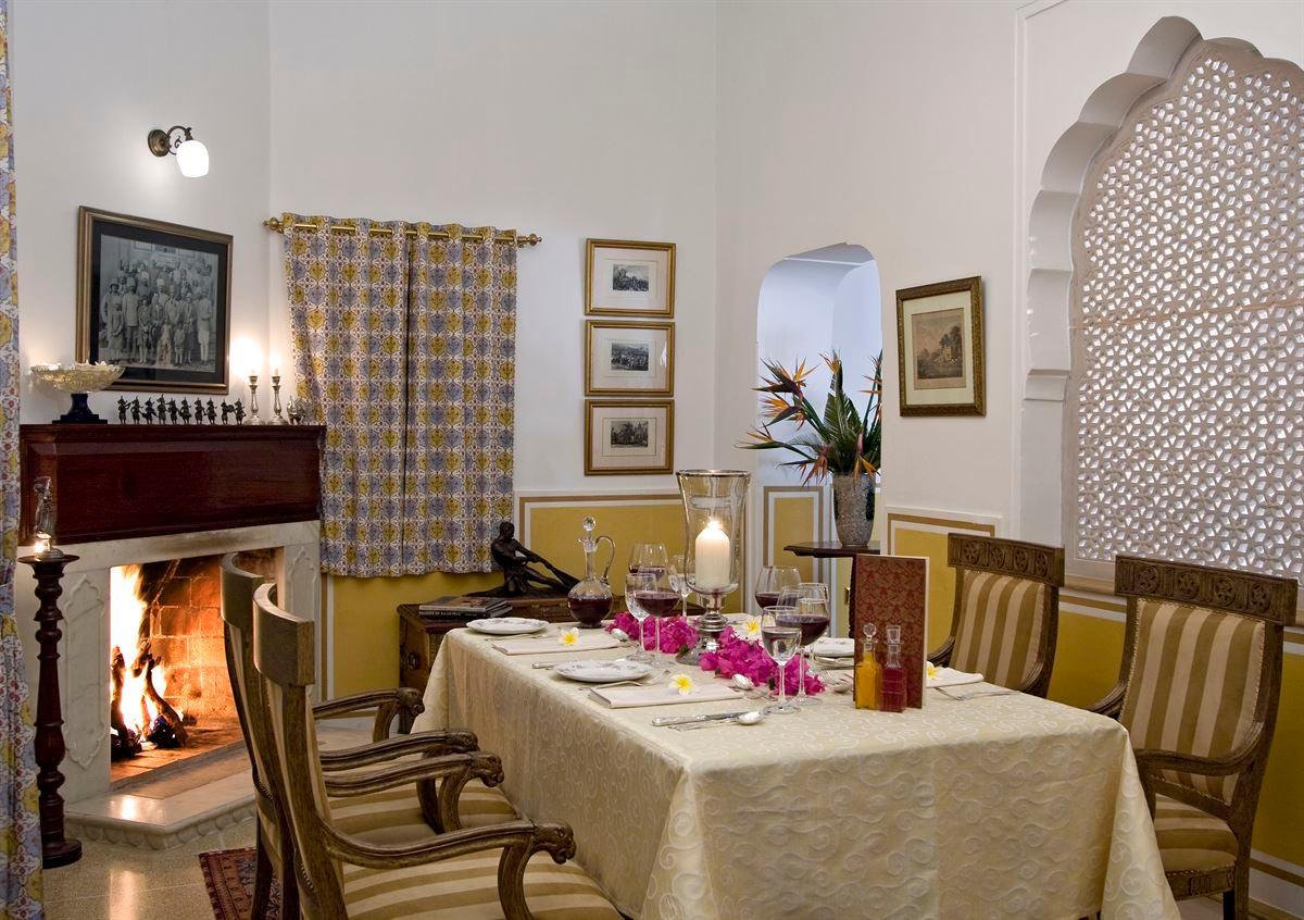 A small dining table set for two with wine glasses and flowers in front of a lit fireplace.