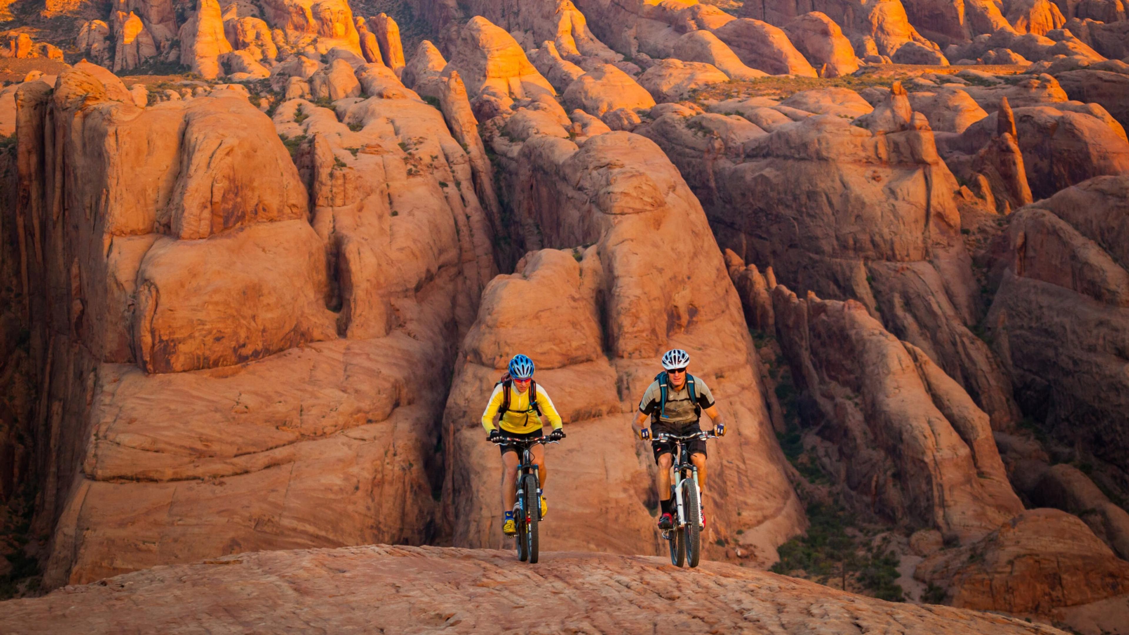 Two cyclists in helmets ride mountain bikes along a ridge of red rock formations.