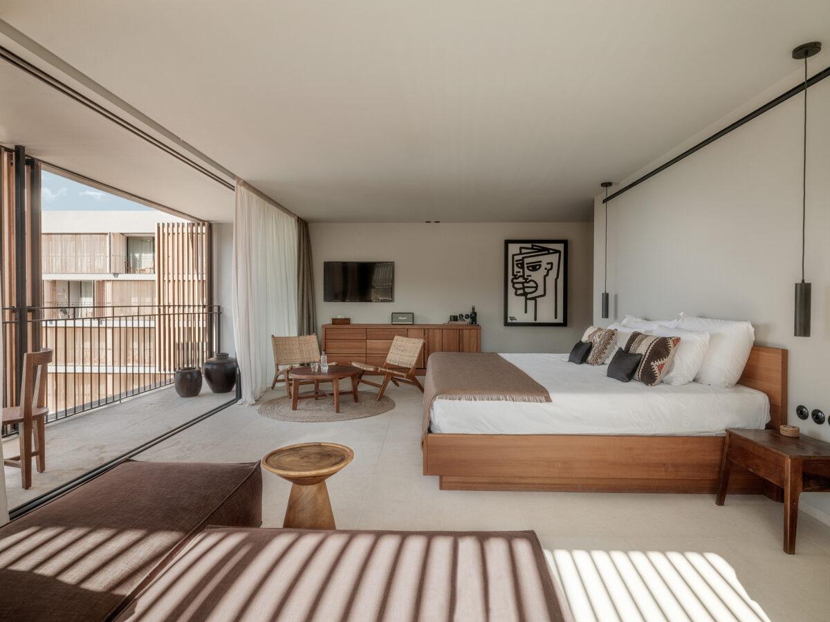 A bedroom with a king bed, a striped rug and a grey sofa in the foreground, and a balcony beyond open glass doors onto a timber-clad building.