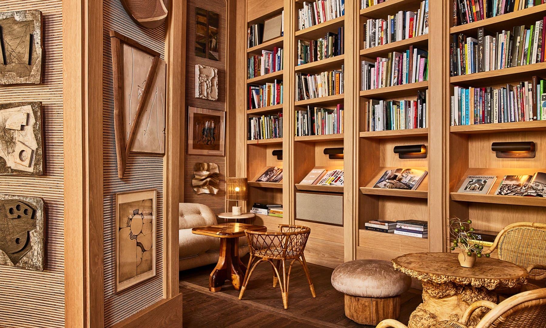 A reading nook lined with bookshelves beside a tiled wall hung with abstract wooden artworks, with a rattan chair and ottoman.