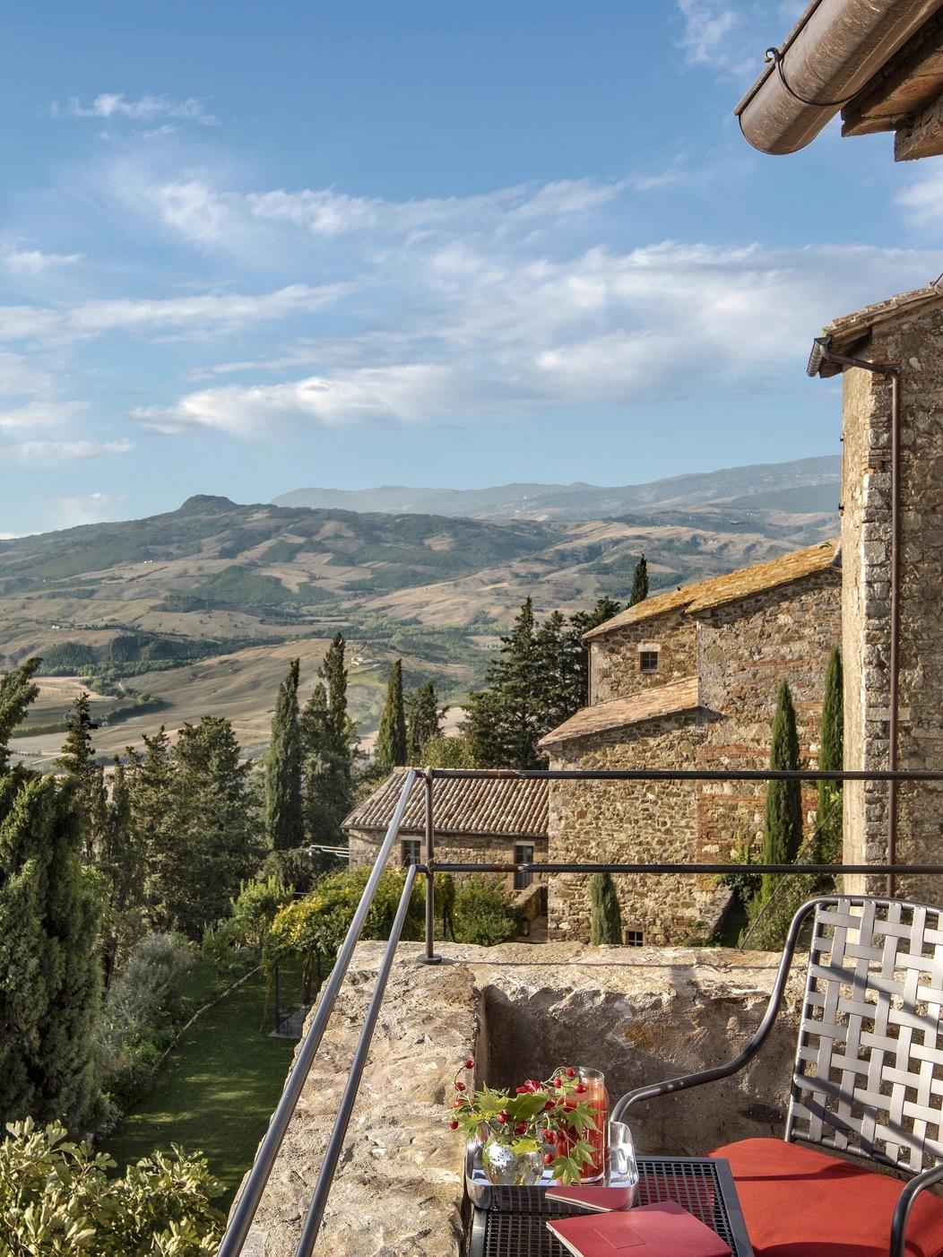 A wrought iron chair and small table with flowers and a drink on a stone terrace, overlooking wooded hills and mountains under a partly clouded sky.