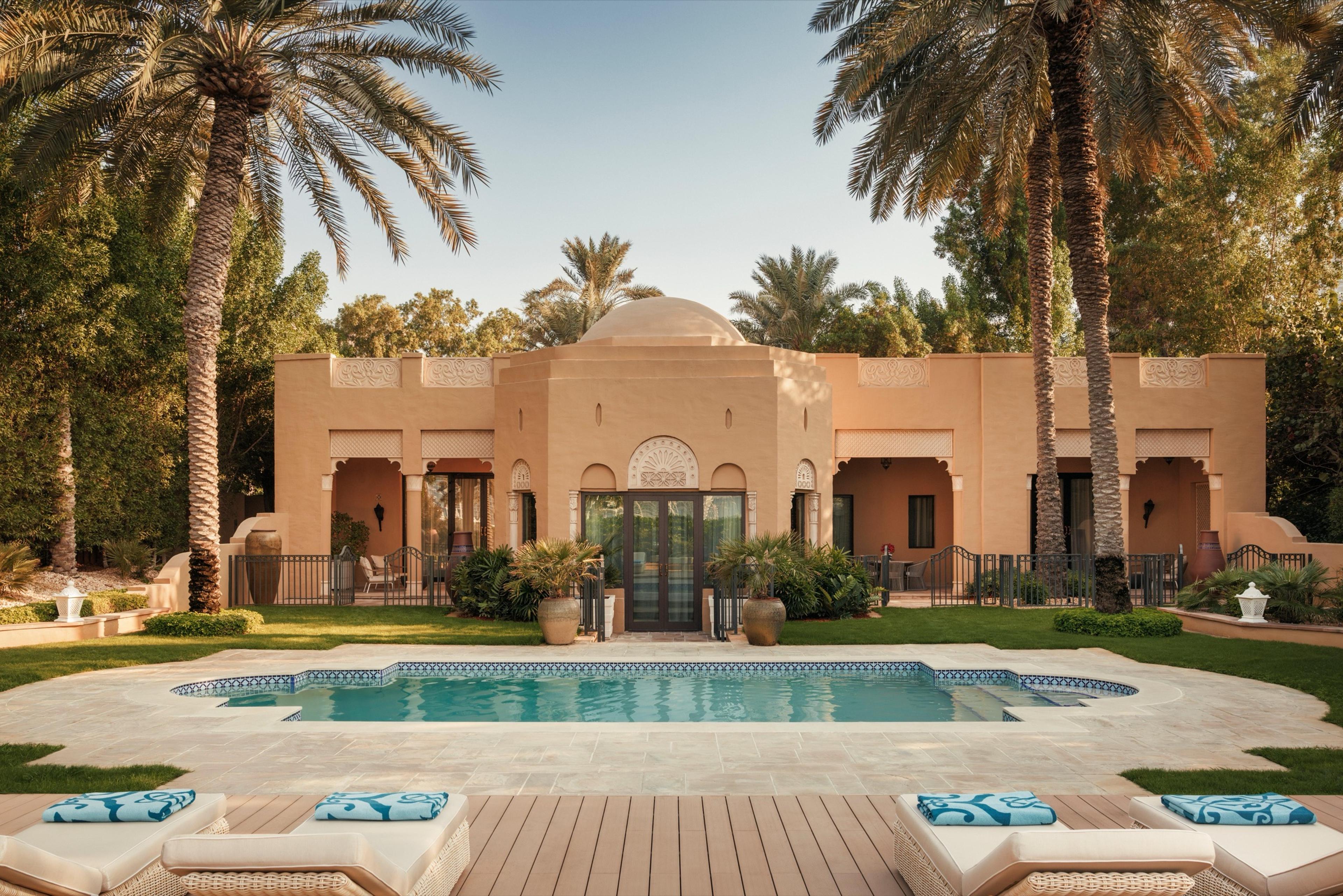 A domed, Moorish-style villa sits behind a curved pool and palm trees, with cushioned loungers in the foreground.