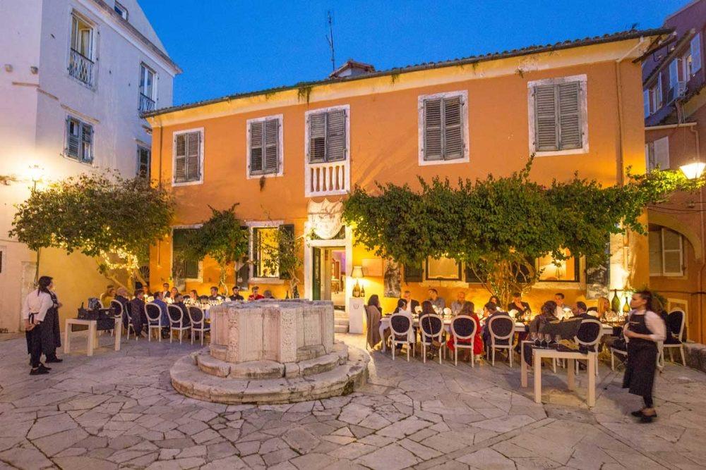 Tables set around an old stone well in a small ochre square at dusk