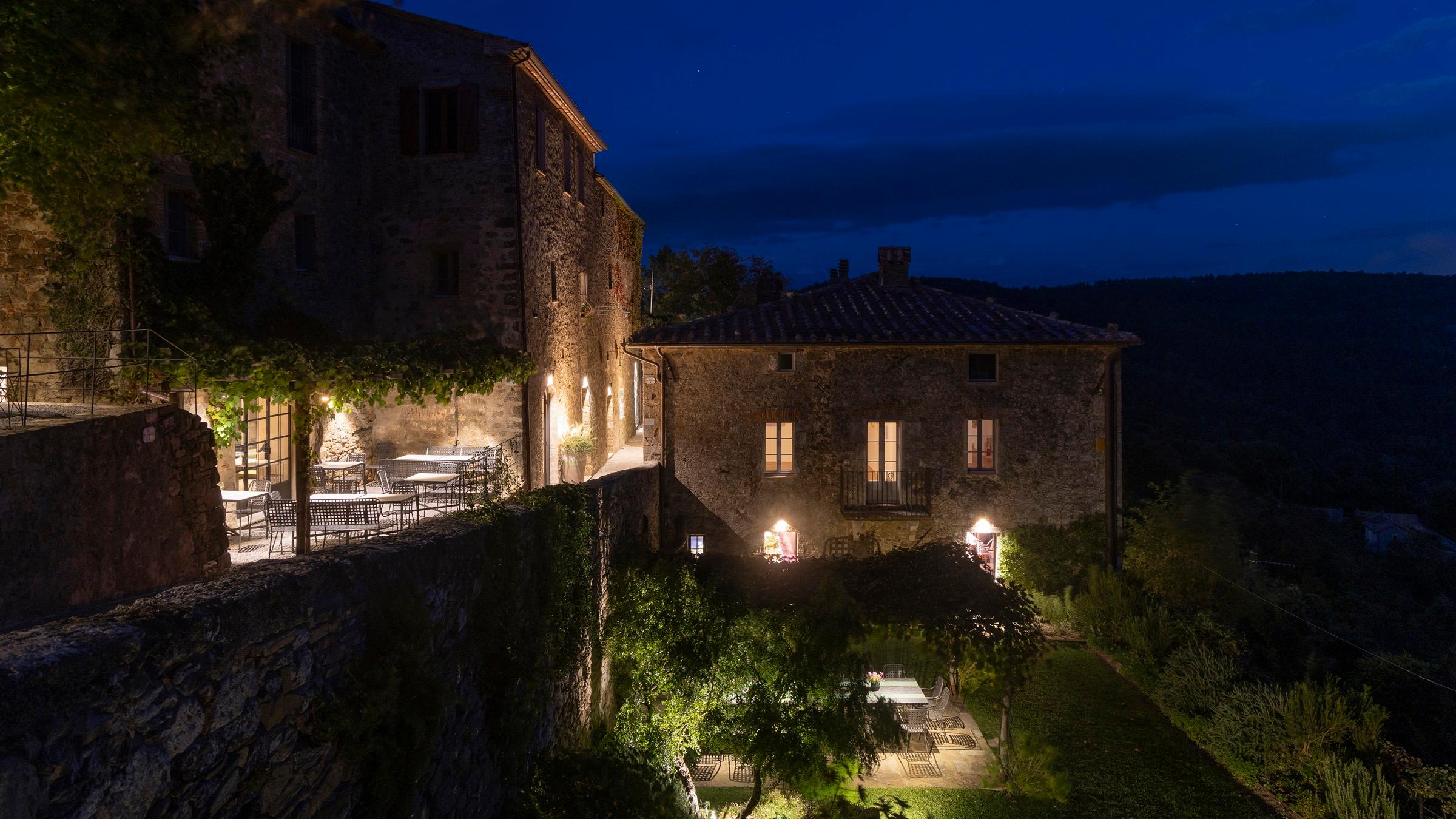 A stone hamlet lit up at night, warm light in the windows and an outdoor dining terrace glowing below, against a deep blue sky.