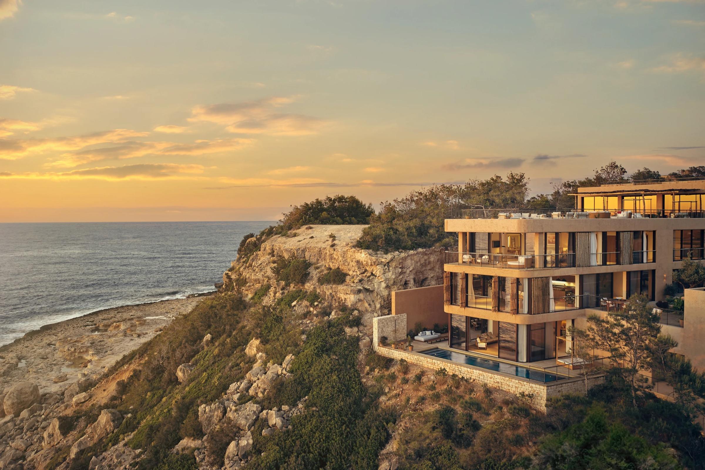 A clifftop villa at sunset, three storeys of glass and pale stone, with a small pool on a terrace below and a rocky shore falling away to the sea on the left.