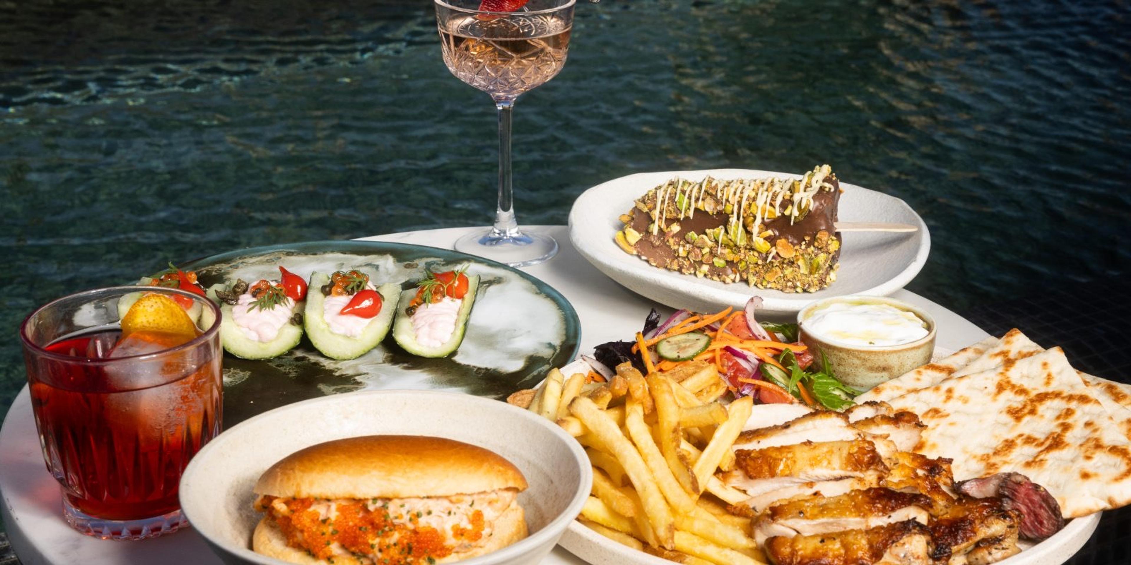 A round poolside table set with a cocktail, a glass of wine, dips, flatbread, a salad, fries, and a burger, with pool water behind.