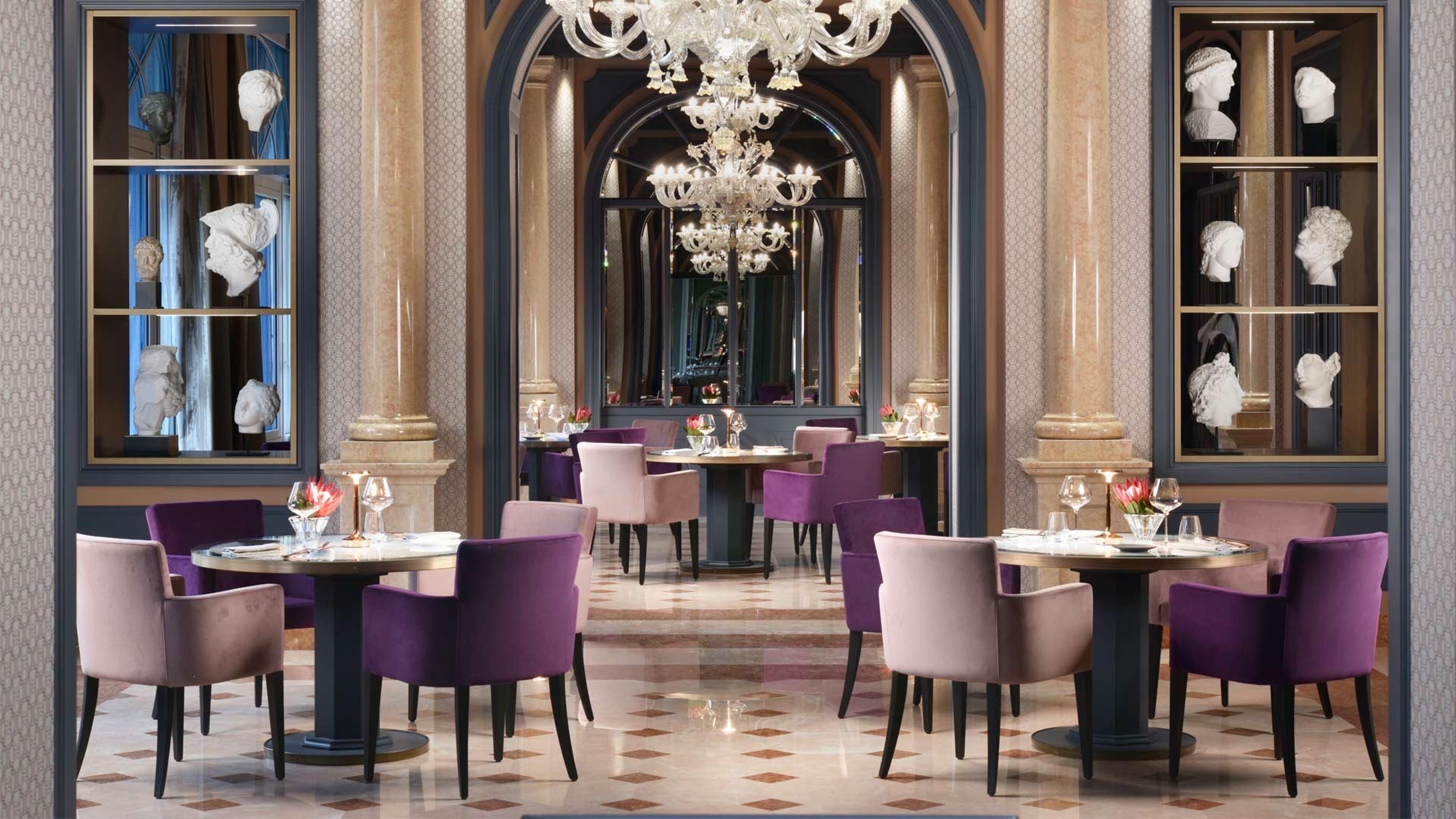 A restaurant interior with purple upholstered chairs around marble-topped tables, columns and classical busts displayed in wall cases.