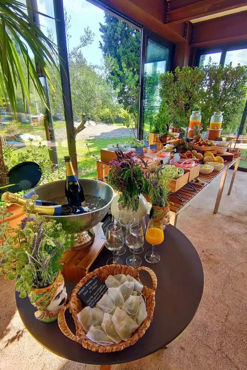 A breakfast table spread with juice dispensers, bowls of fruit and pastries, a champagne bucket and a basket of tea bags.