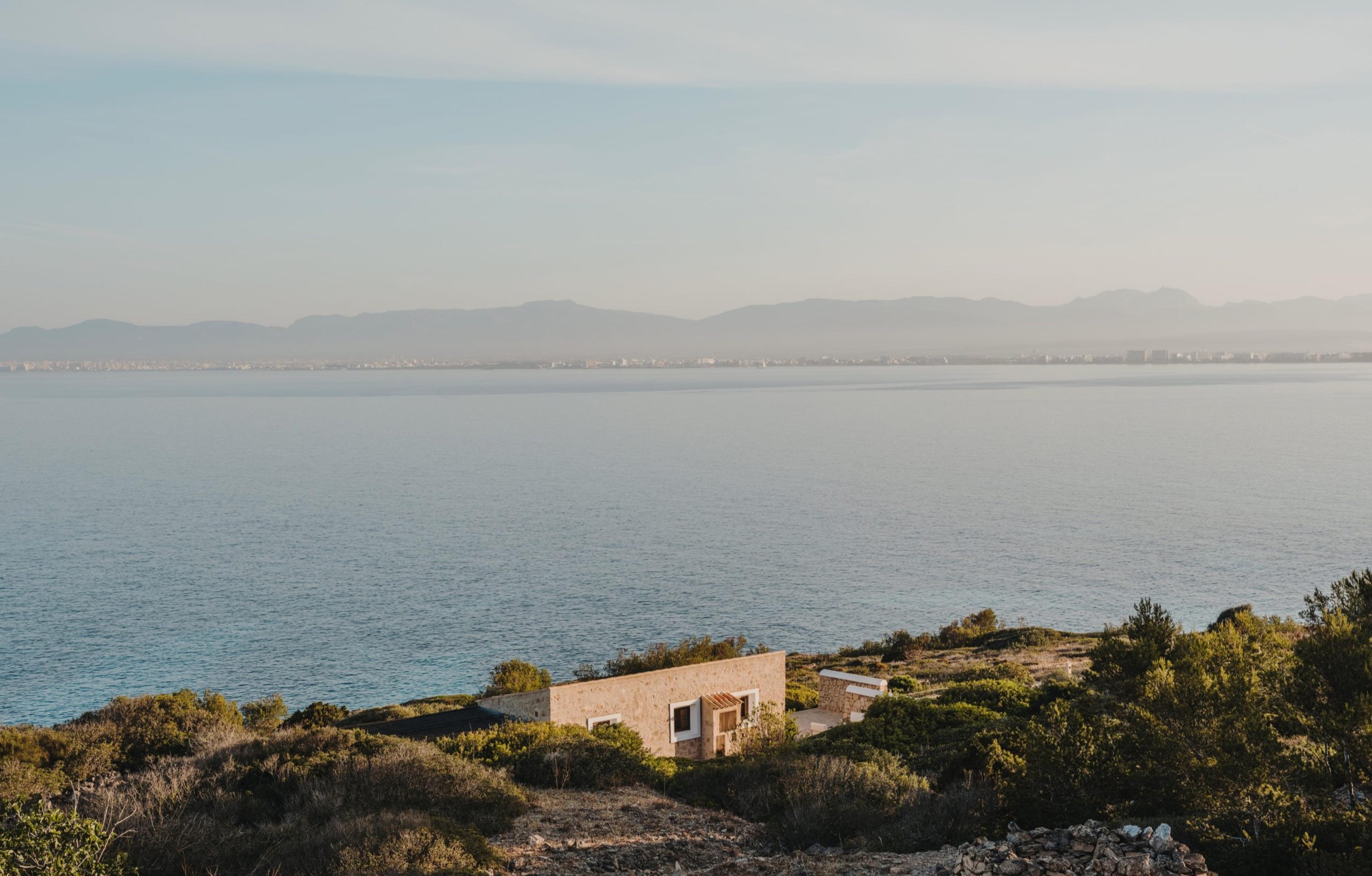 A small stone building on a headland above the sea, seen from a distance with hills and a hazy coastline beyond.