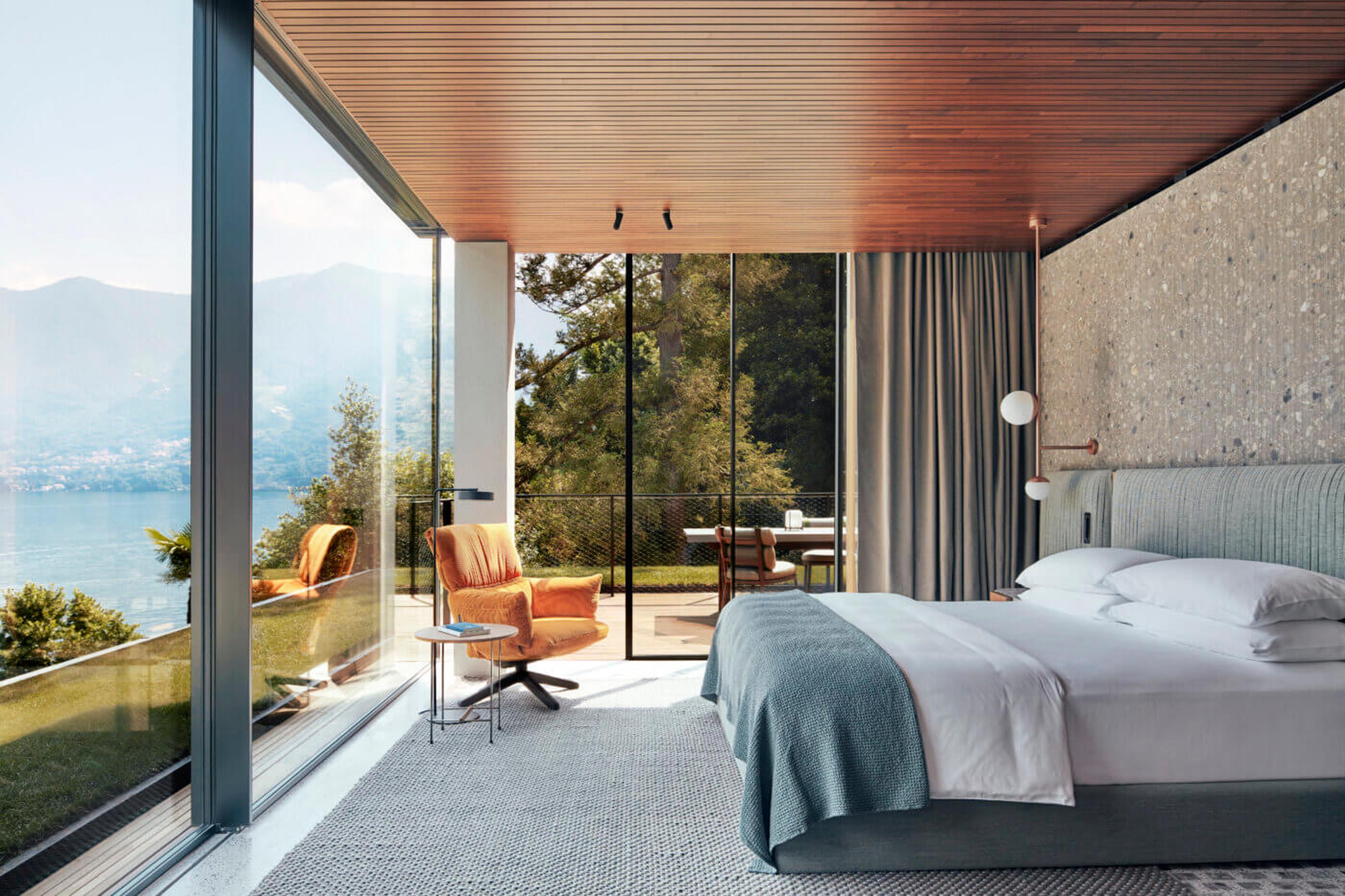A hotel bedroom with orange lounge chairs, glass walls on two sides, and a view through trees to a lakeside dining terrace.
