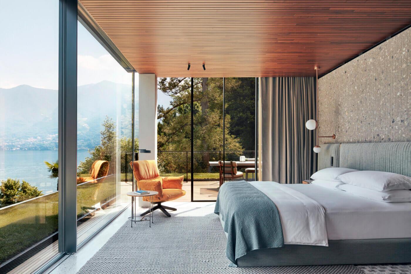 A hotel bedroom with orange lounge chairs, glass walls on two sides, and a view through trees to a lakeside dining terrace.