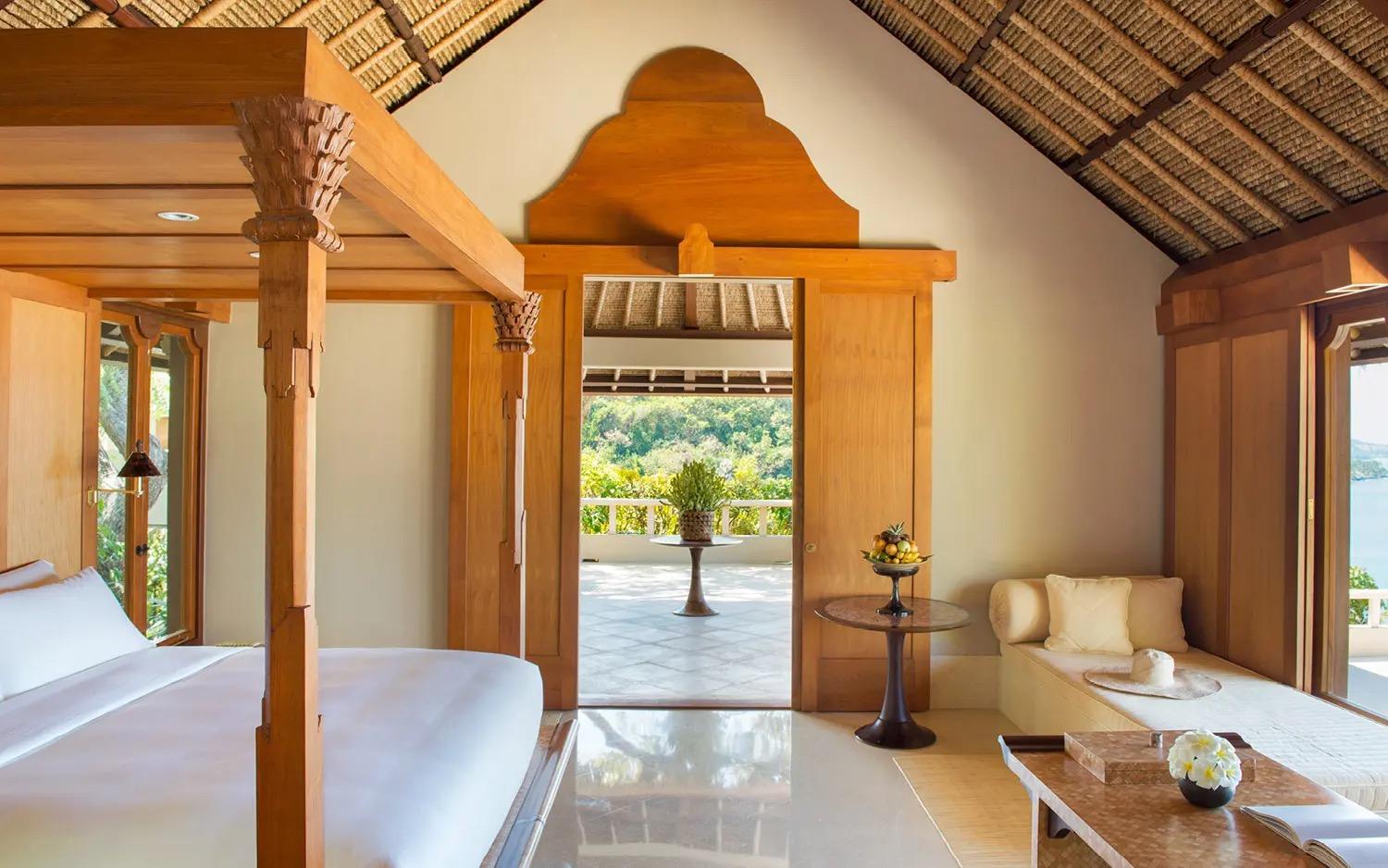 A bedroom with a four-poster wooden bed and a daybed, its door open onto a terrace with trees and water beyond.