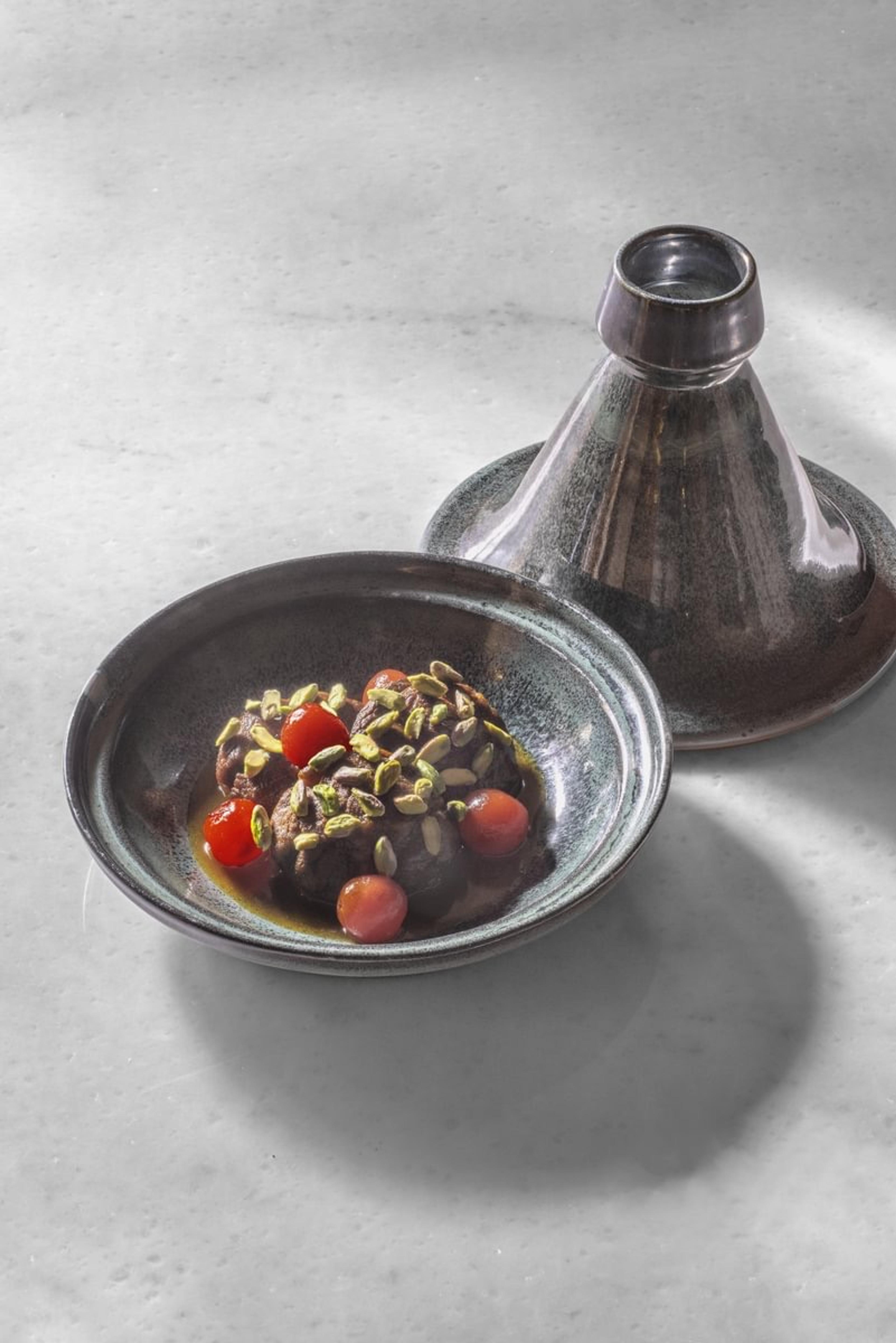 A tagine dish of meatballs with cherry tomatoes and chopped pistachios, the conical lid set beside it on a stone surface.