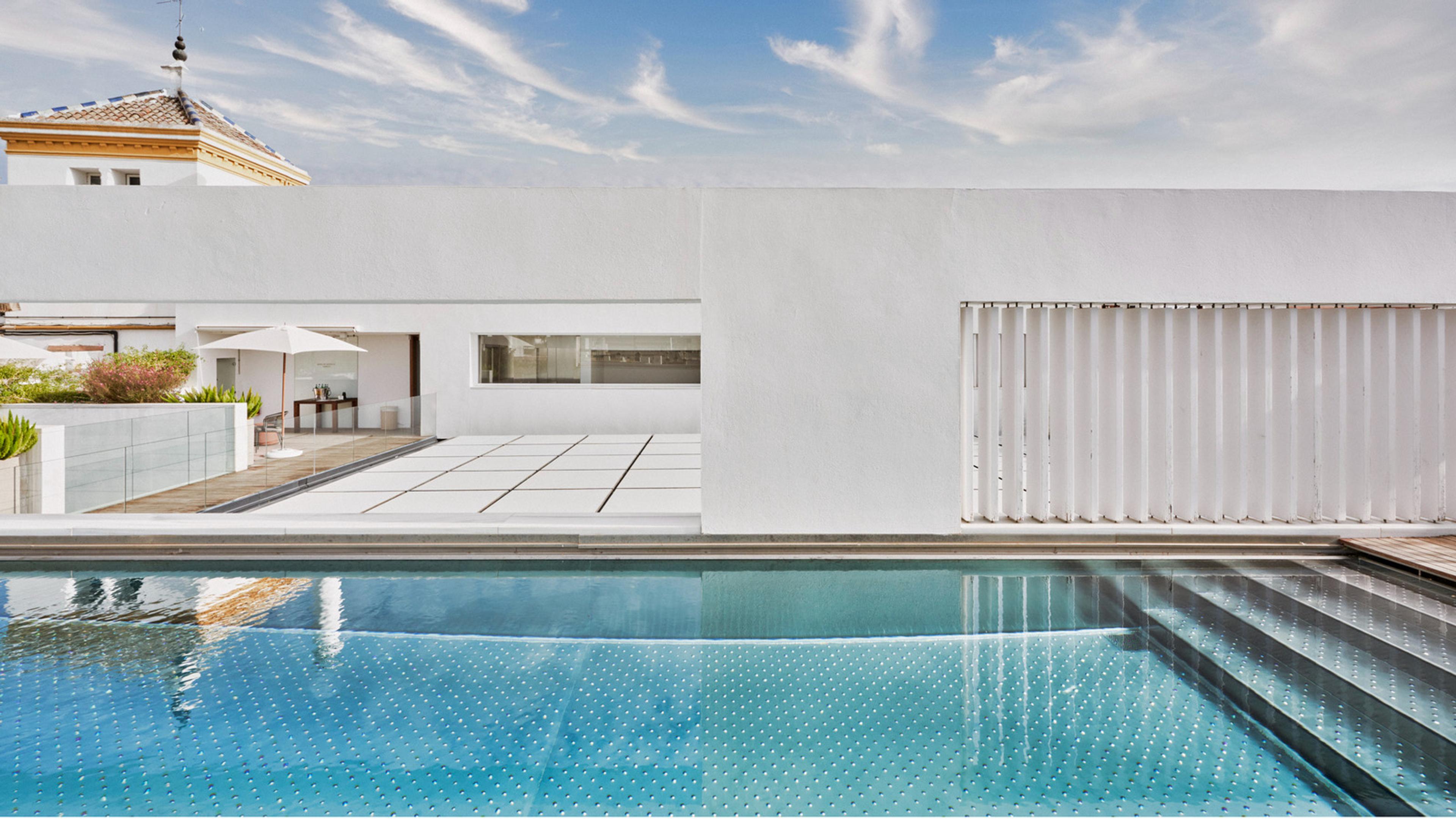 A long rooftop swimming pool reflects a pale blue sky, with a white terrace, a folded umbrella, and a tiled church tower visible beyond the building's edge.