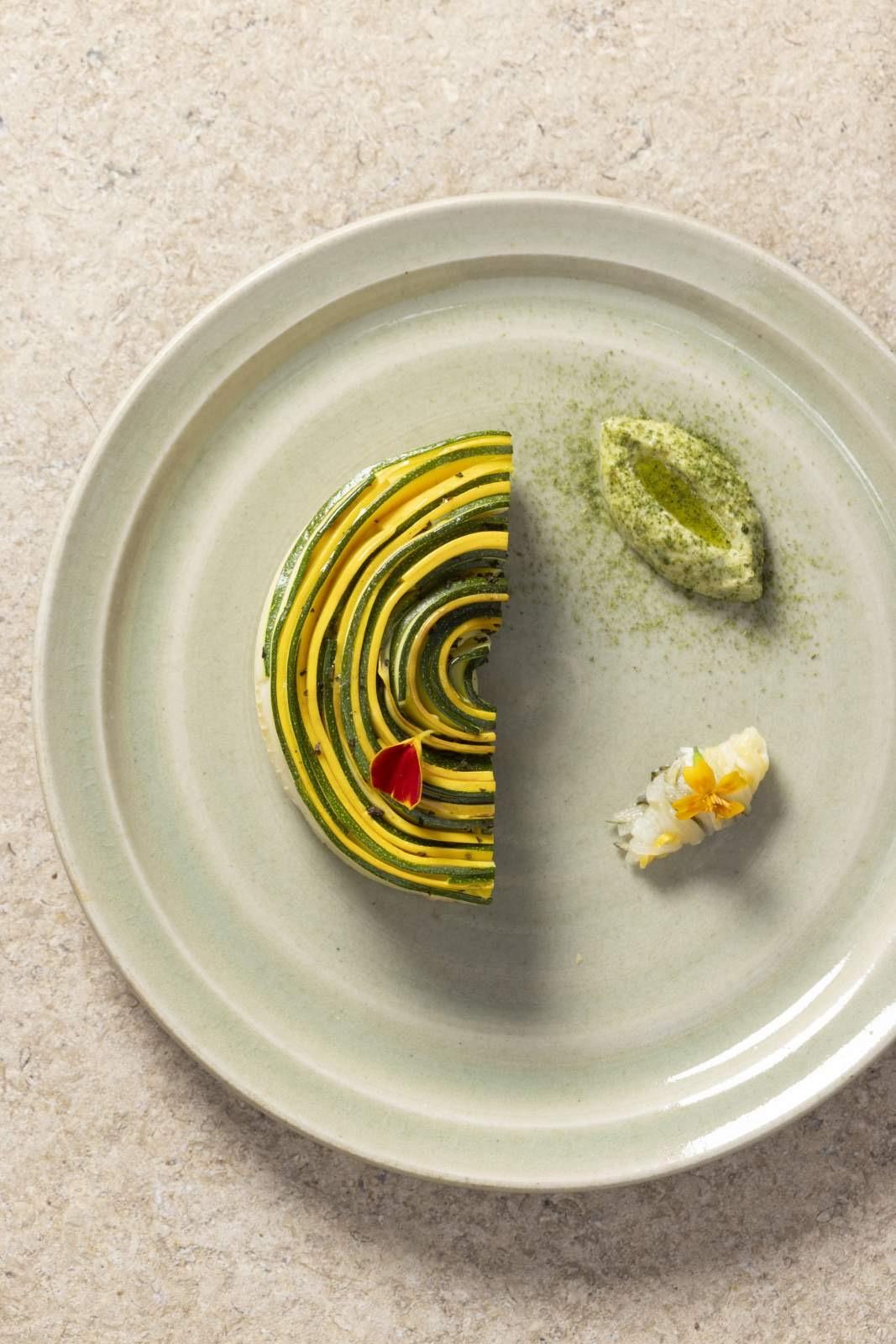 A plated dish of layered courgette rounds with a quenelle of green sauce and a small garnish, on a pale ceramic plate.
