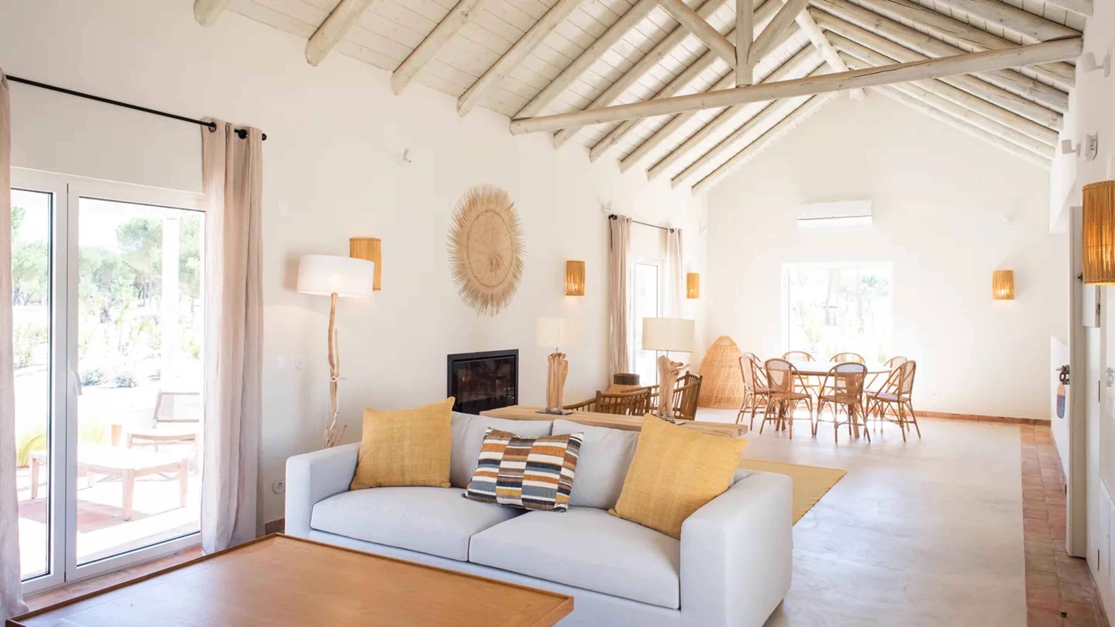 a living room with a vaulted wood-beamed ceiling, a pale sofa with mustard cushions, and a dining table in the background