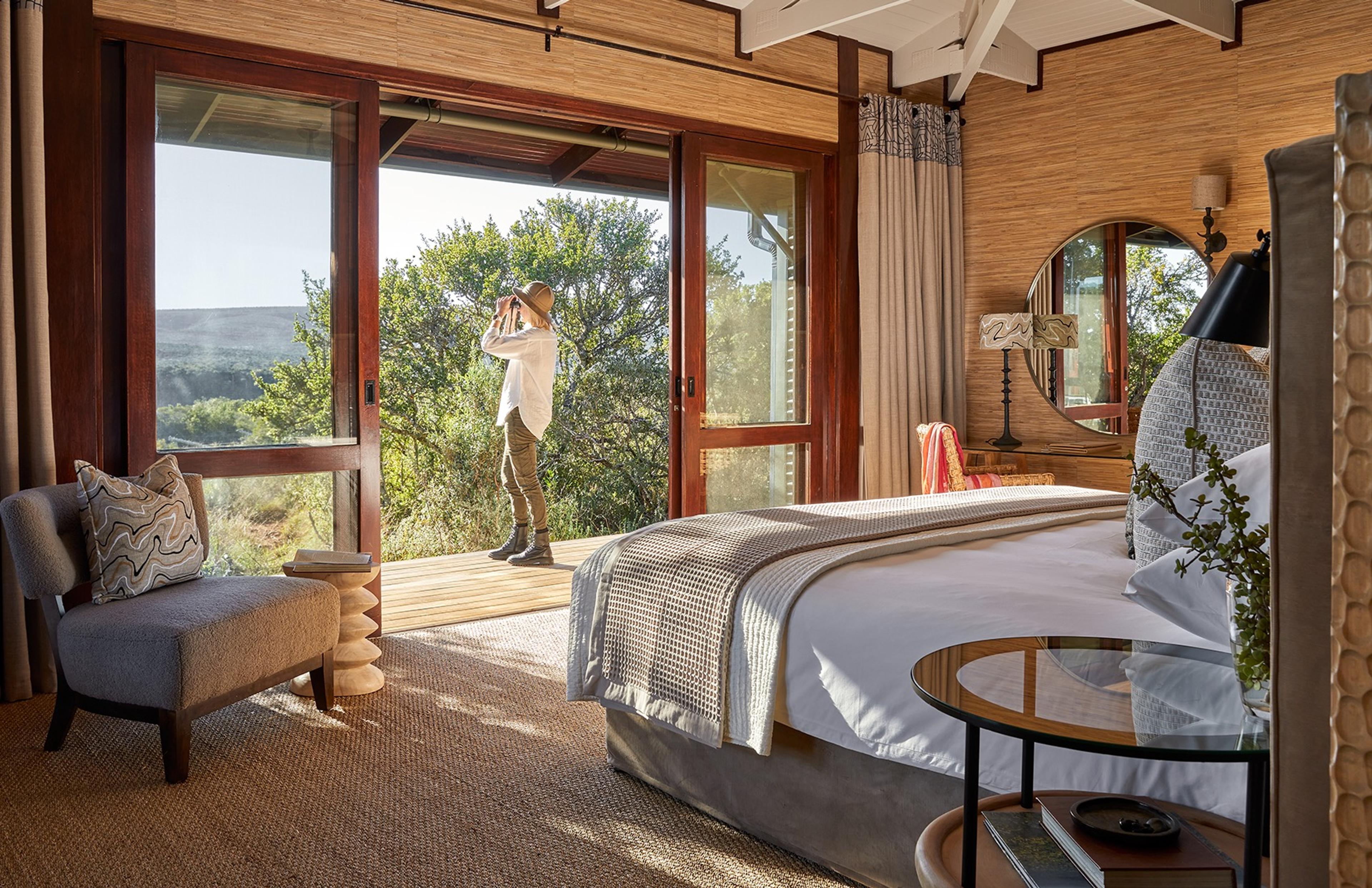 A bedroom with a made bed and a glass-topped side table, a person standing on the deck outside looking through binoculars.