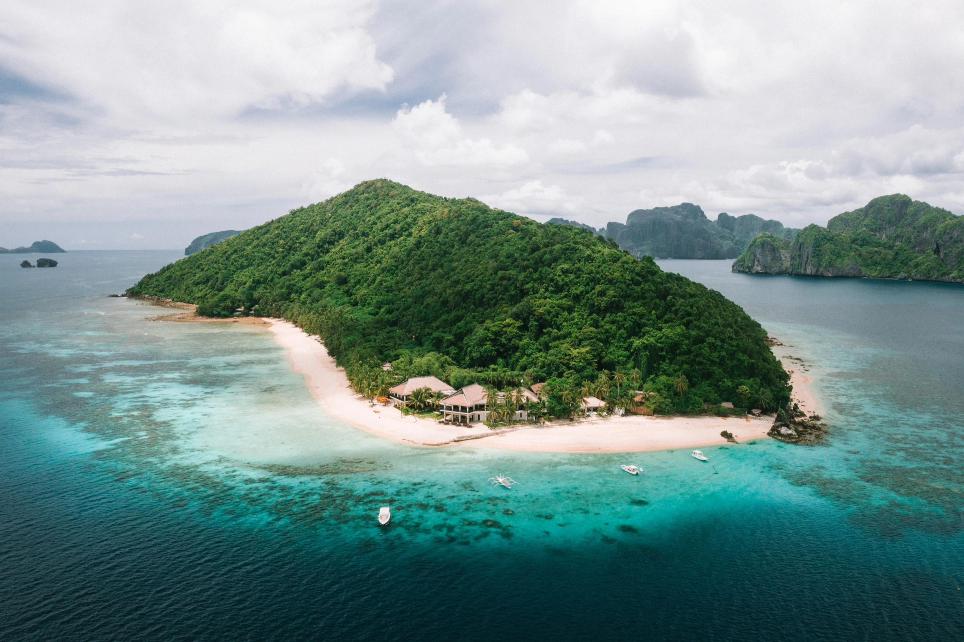 An aerial view of a green island with a long white beach and pale shallows, more islands on the horizon.