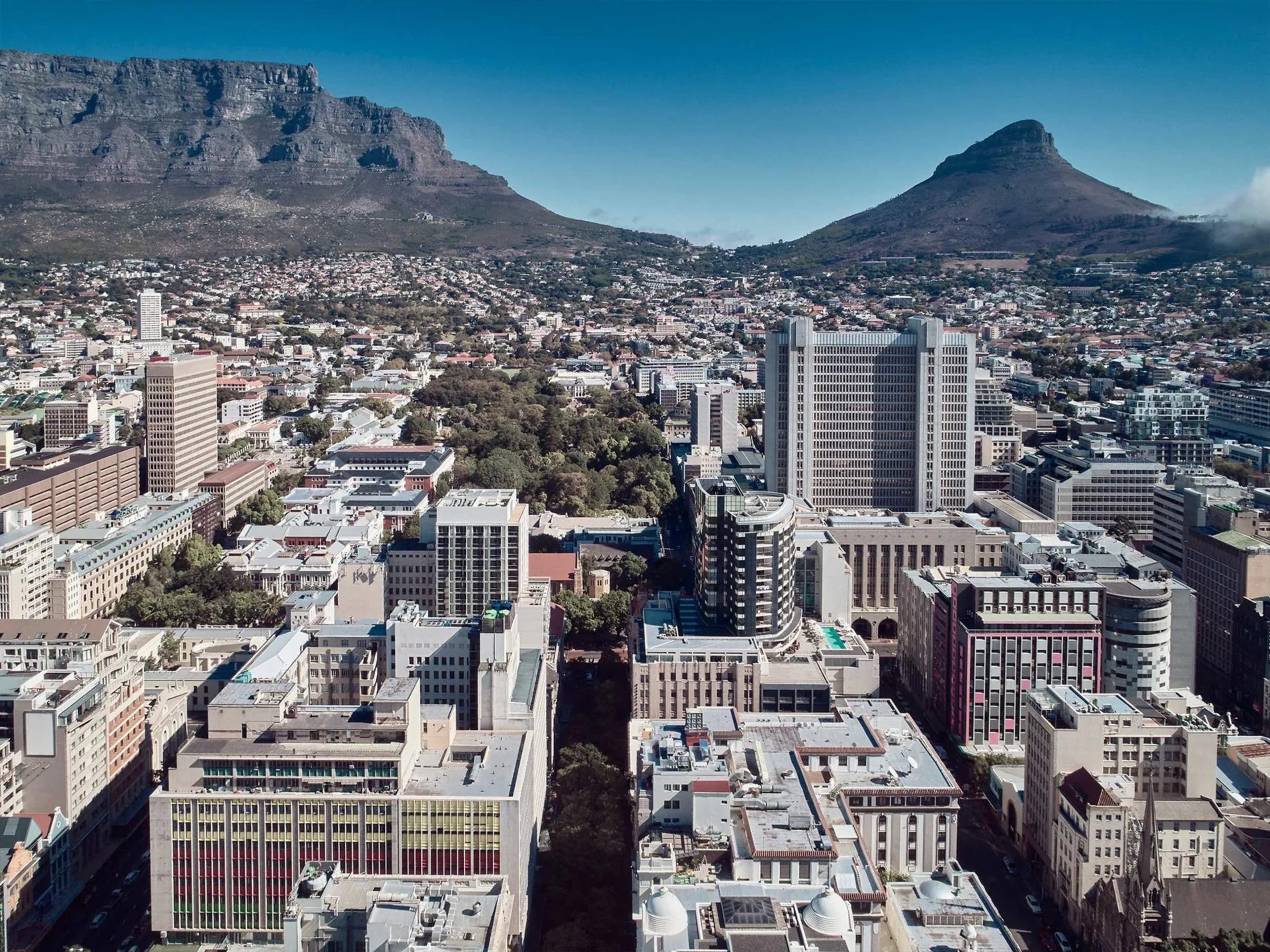 An aerial view over a dense city centre of tall office blocks and tree-lined streets, with Table Mountain and Lion's Head on the skyline under a clear blue sky.