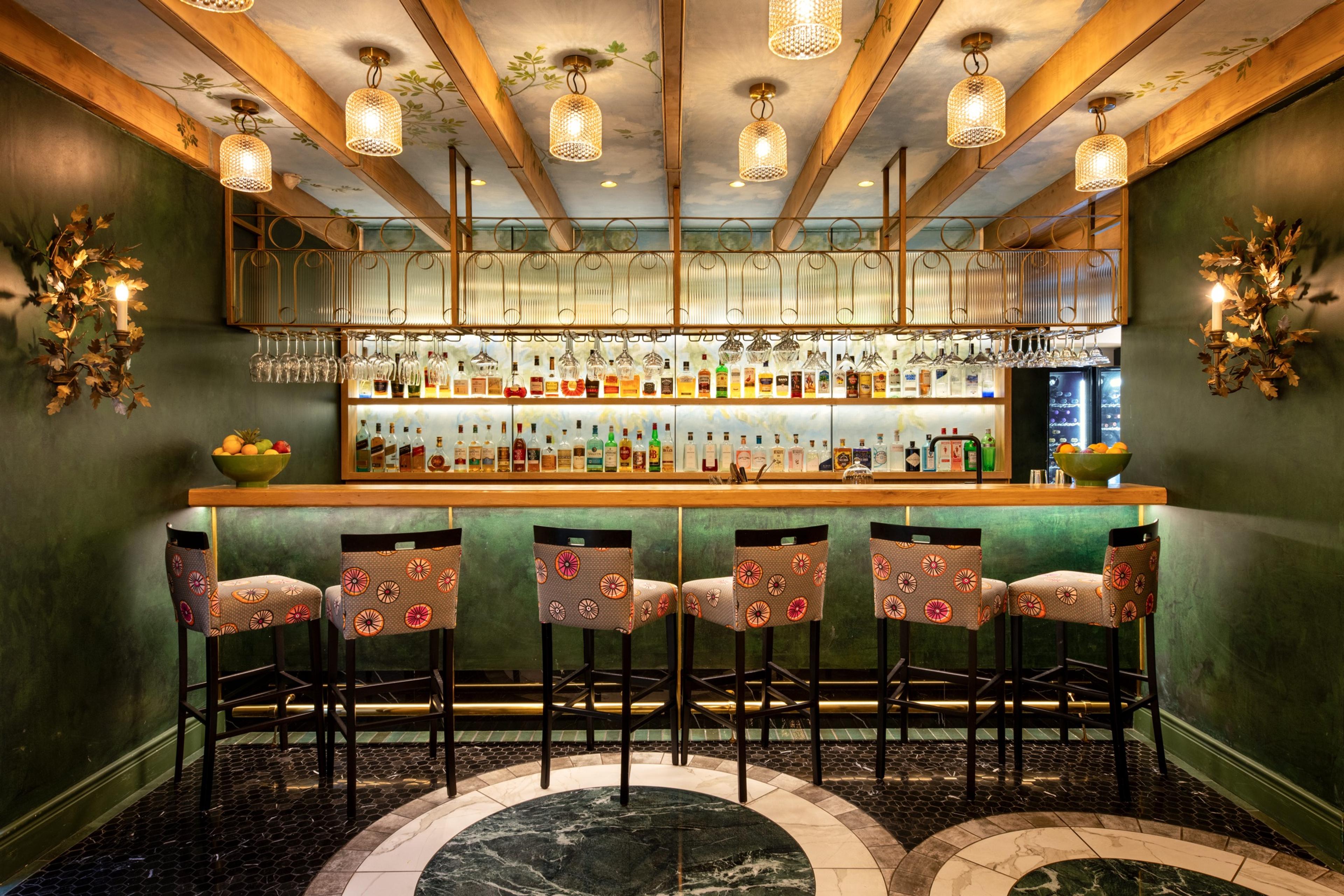 A green-walled bar with a row of upholstered stools, glasses hanging above the counter and a dark floor with a marble inlay.