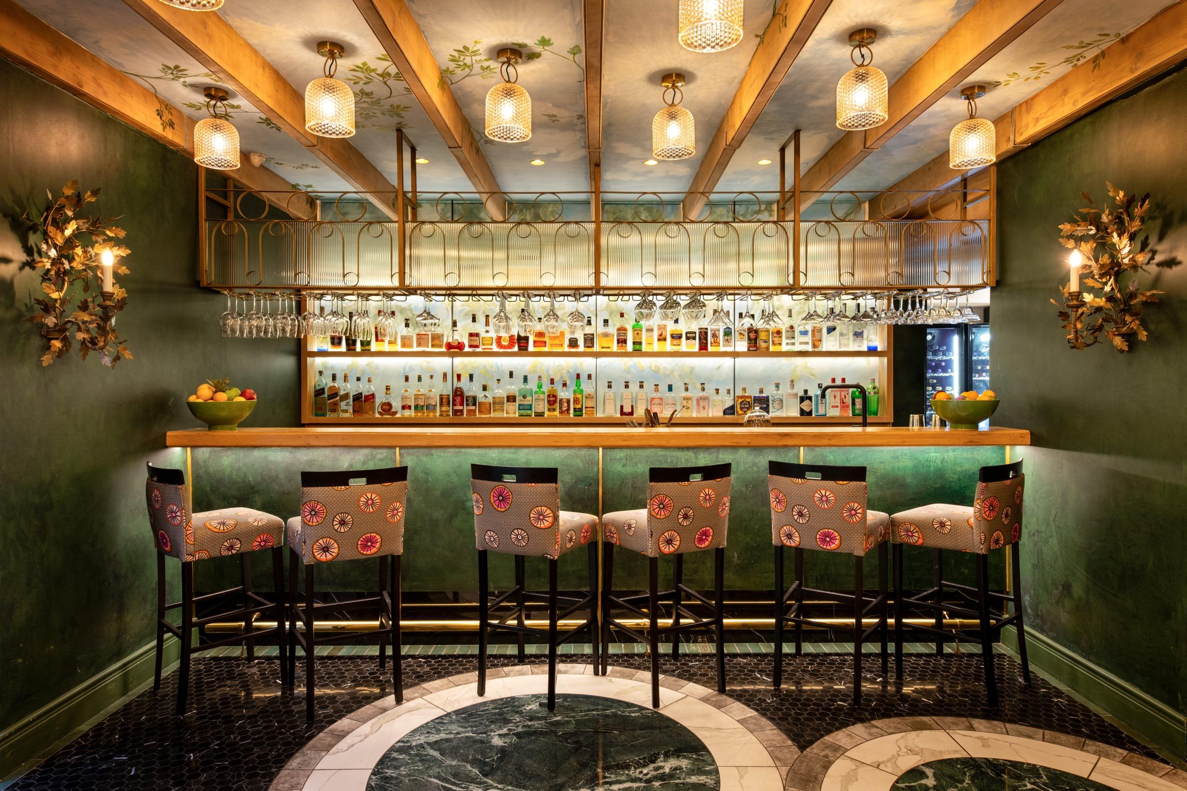 A green-walled bar with a row of upholstered stools, glasses hanging above the counter and a dark floor with a marble inlay.
