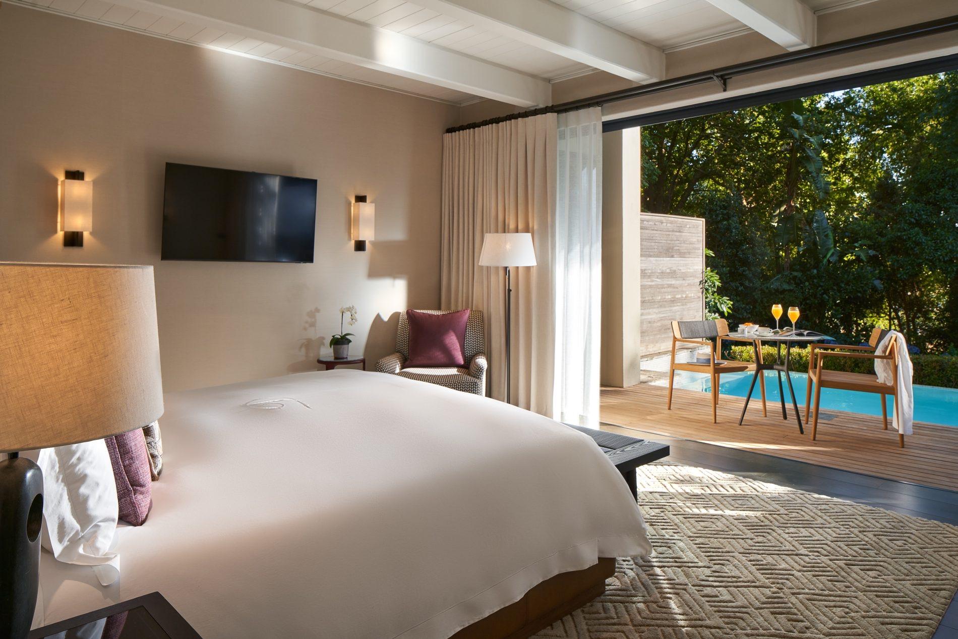 A bedroom with a white bed faces an open glass wall onto a wooden deck, a pool and a dining table beyond.