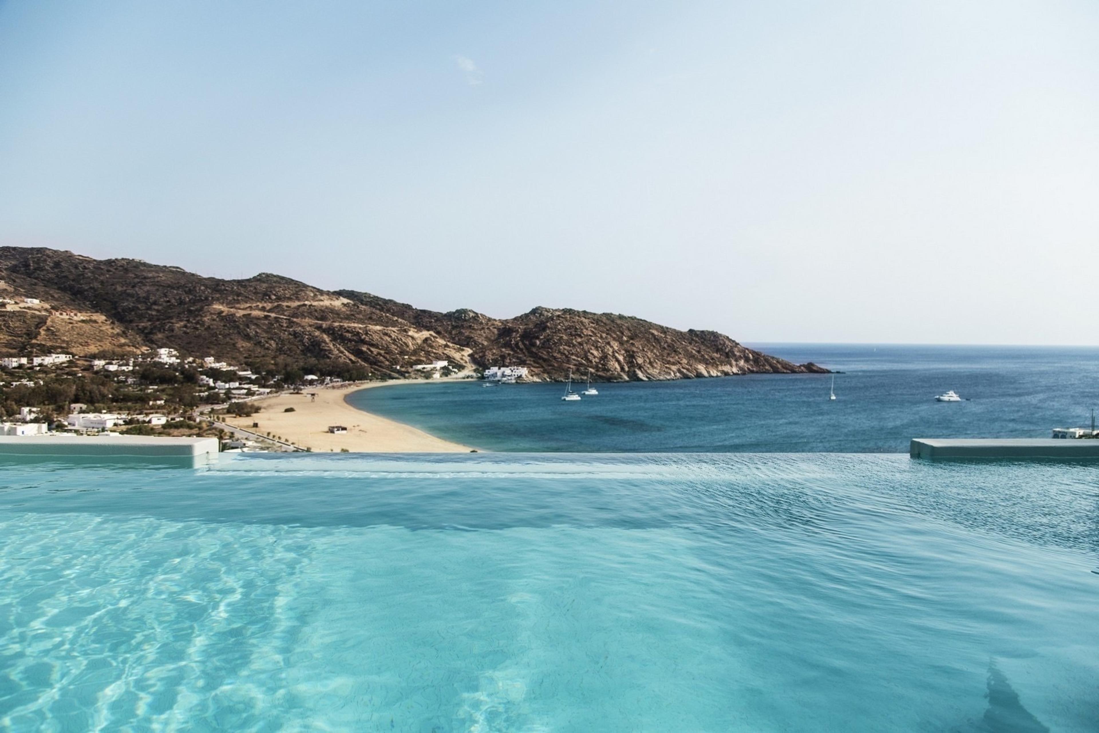 An infinity pool in the foreground, with a sandy bay, white houses on the hillside and small boats on blue water beyond.