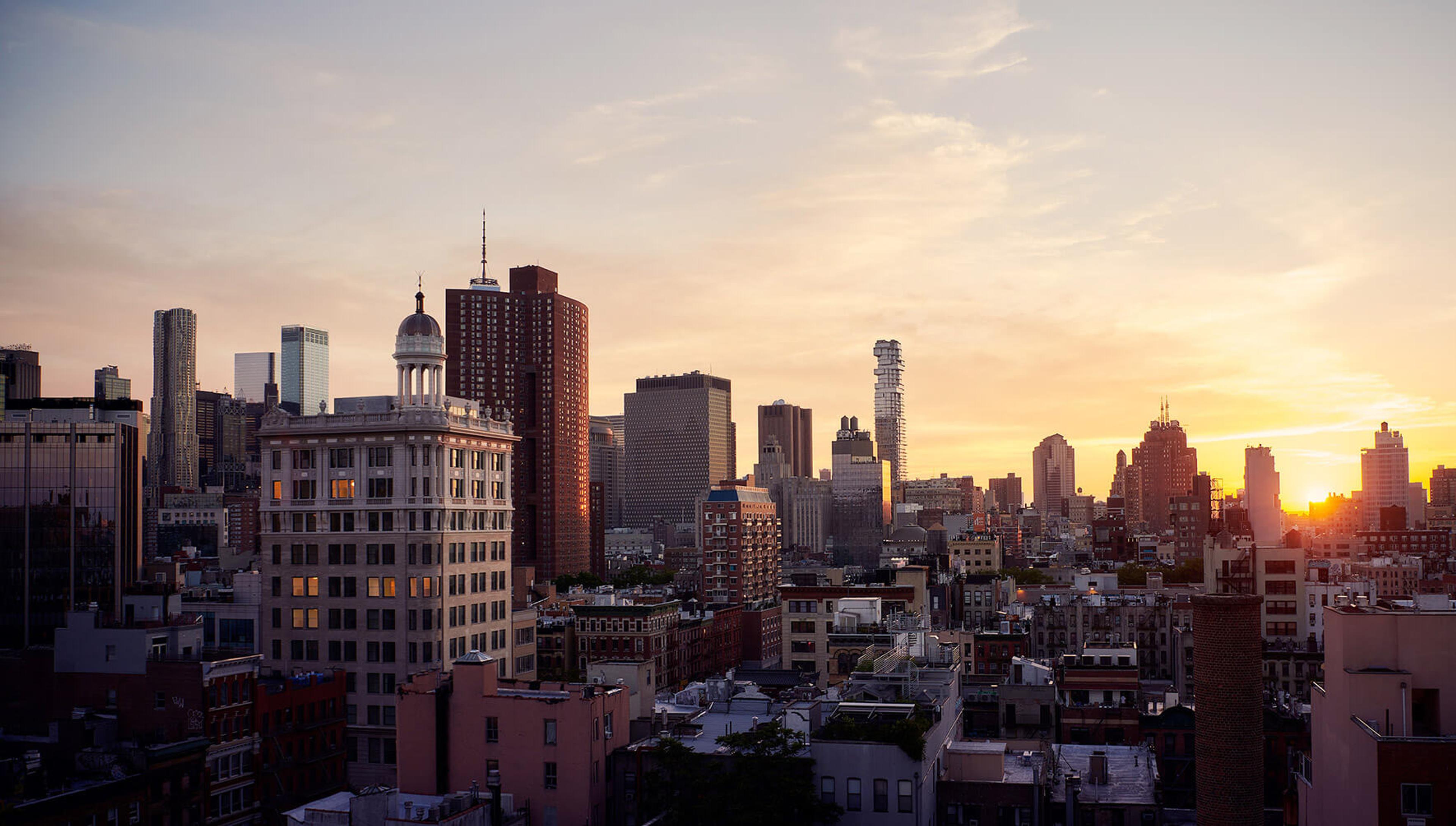 A dusk view over a city skyline of towers and rooftops, with a domed tower in the middle distance and the sun setting on the horizon.