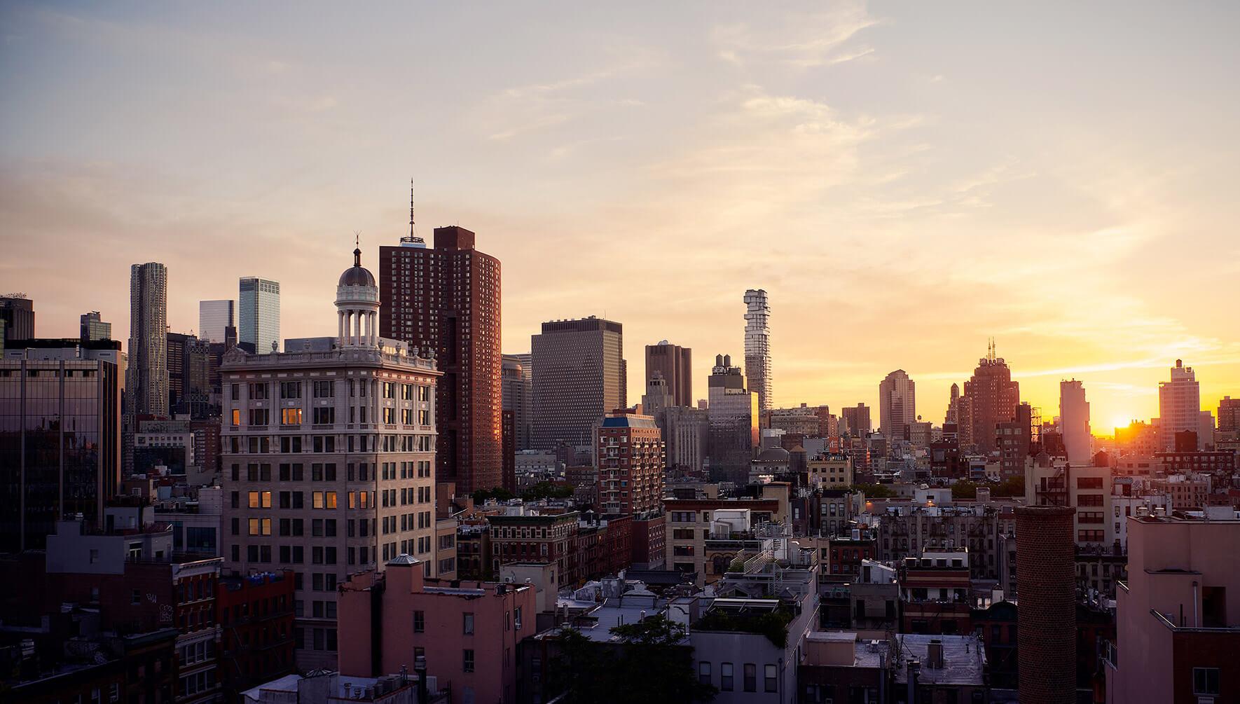 A dusk view over a city skyline of towers and rooftops, with a domed tower in the middle distance and the sun setting on the horizon.