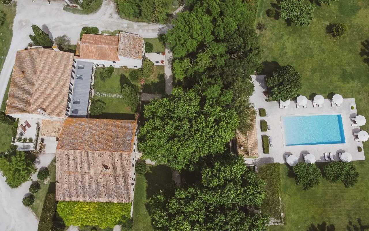 An aerial view of a stone farmhouse complex with terracotta roofs, a rectangular swimming pool and white loungers, surrounded by trees and lawn.