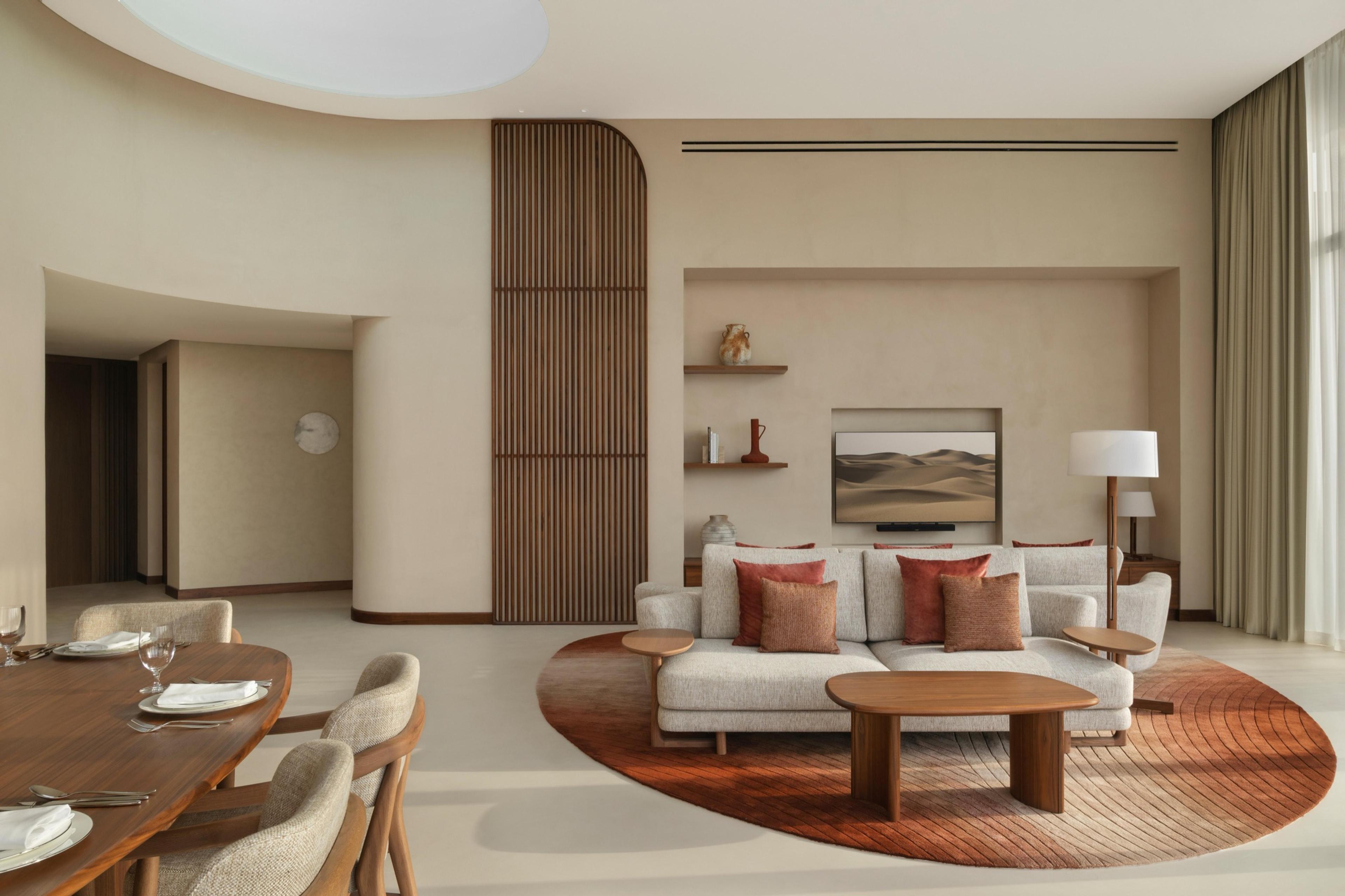 A hotel living and dining room with a round sofa on a red rug, a wood-slat partition, and a dining table set for four in the foreground.