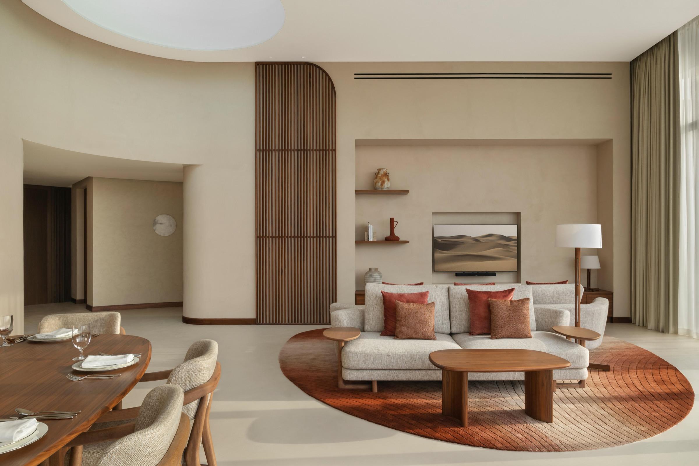 A hotel living and dining room with a round sofa on a red rug, a wood-slat partition, and a dining table set for four in the foreground.