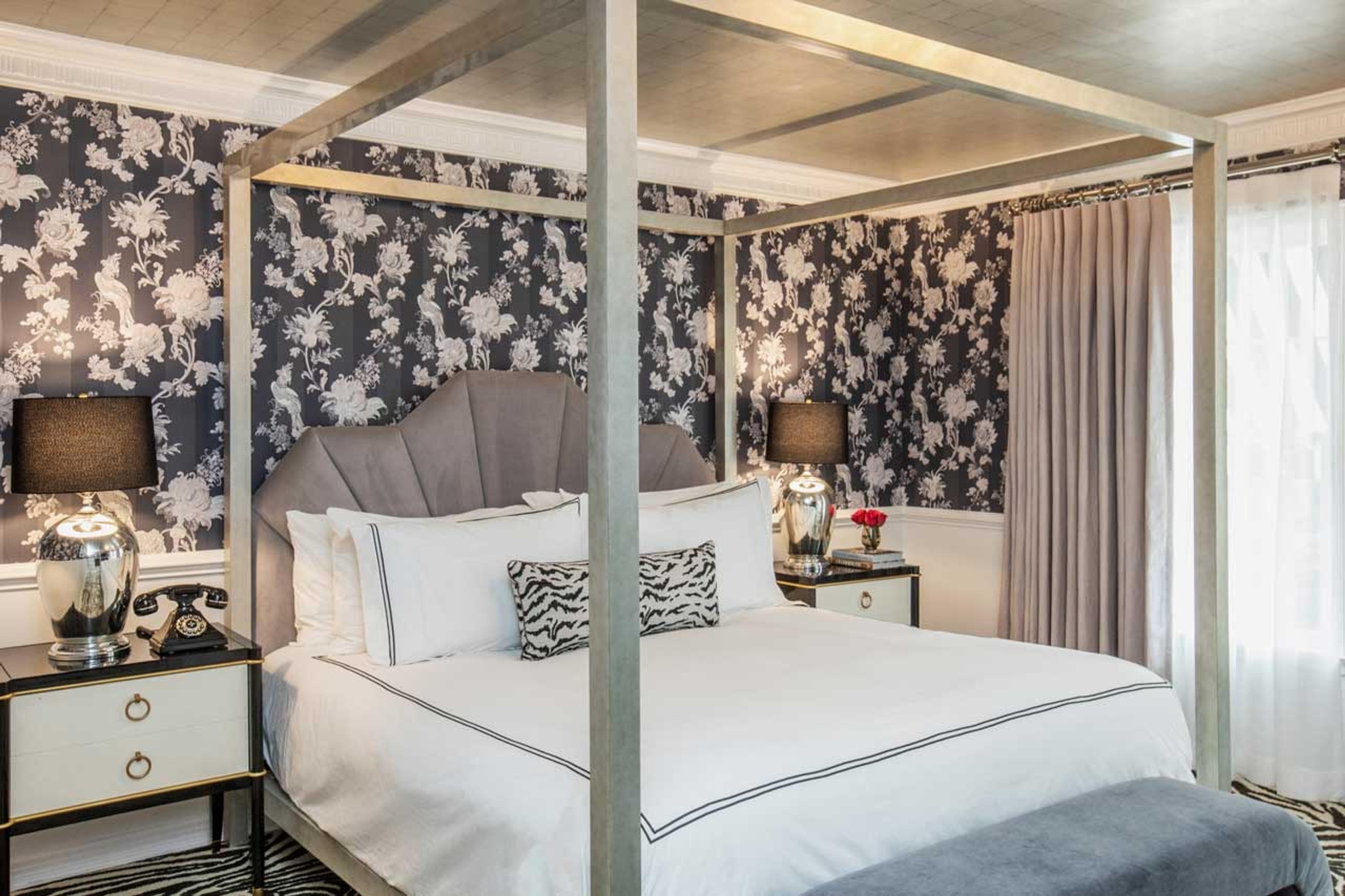 A bedroom with a four poster brass frame bed, a scalloped grey velvet headboard, navy and white floral wallpaper, and a striped zebra print cushion.