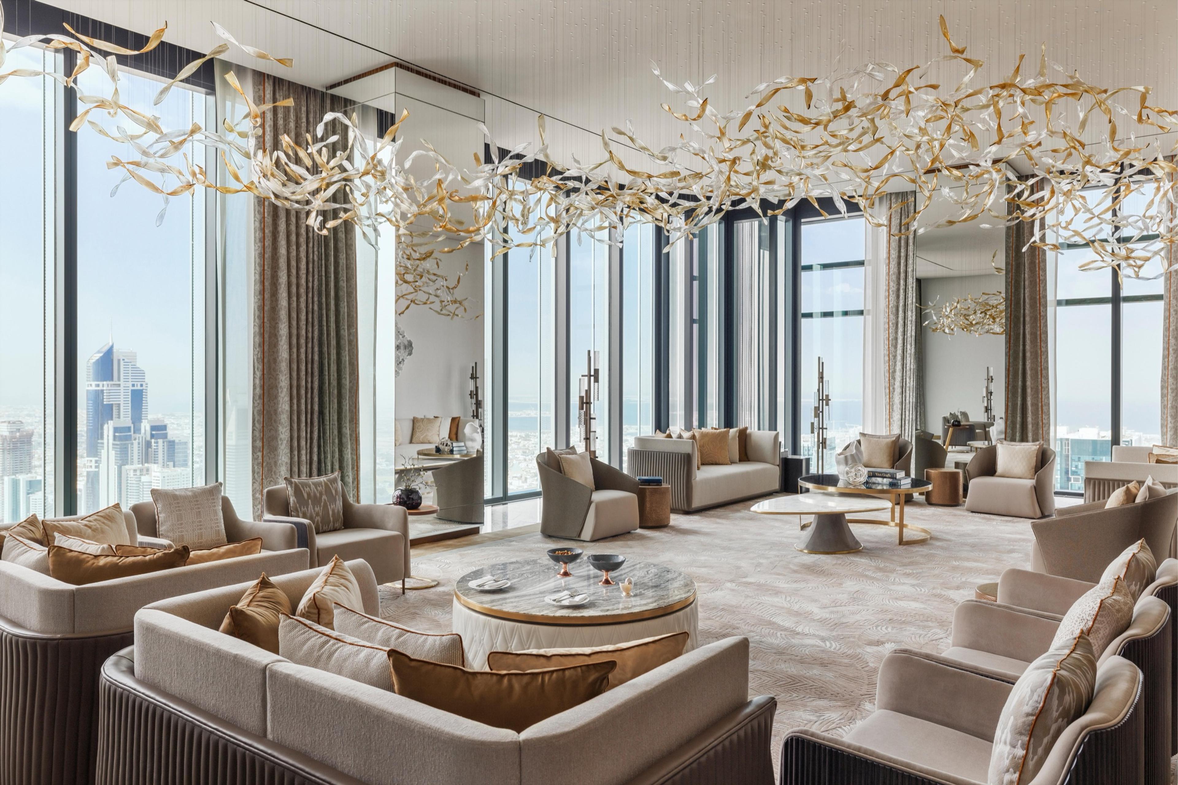 A lounge with a sculptural gold and glass branch chandelier strung across the ceiling, rows of cream sofas and armchairs, and a wall of windows overlooking the sea and city.