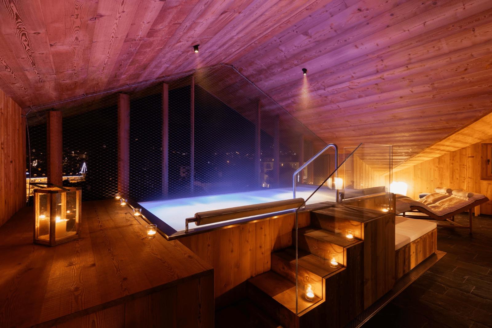 A lit outdoor plunge pool steams at night beside candlelit steps, with a town's lights visible beyond the timber deck.