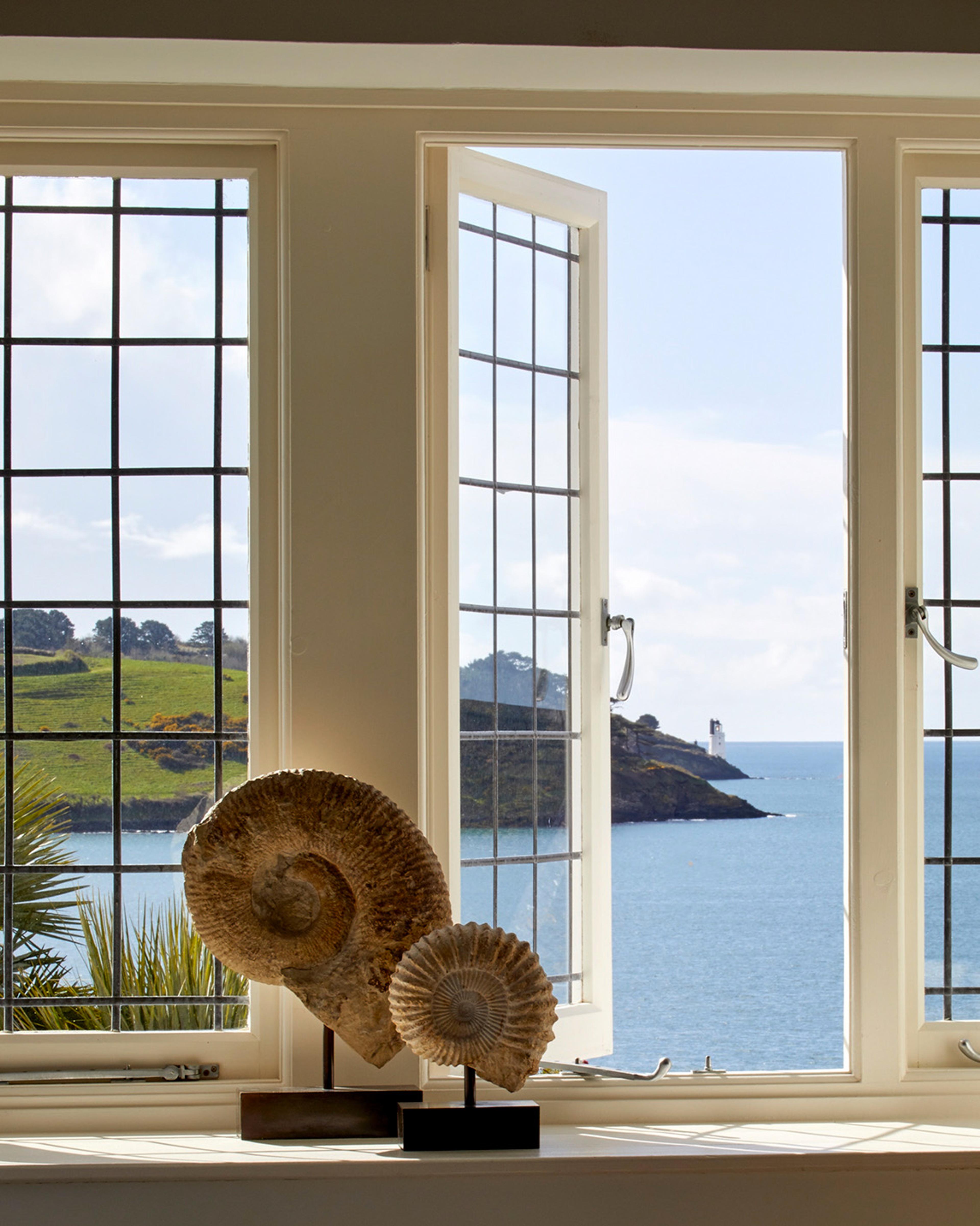 Two large fossil ammonites on stands on a windowsill, with leaded windows open onto a view of the sea and a headland with a lighthouse.