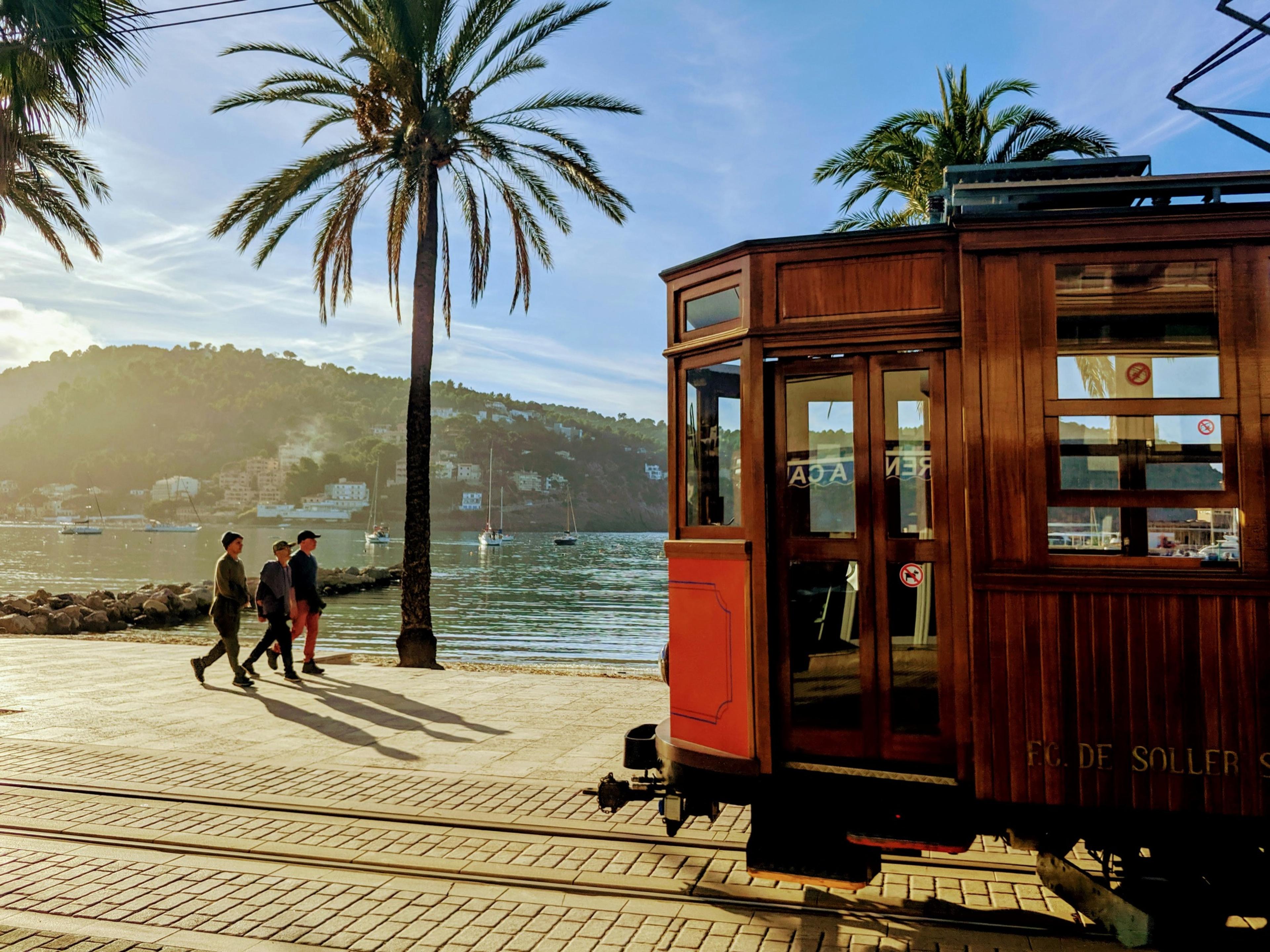 An old wooden tram with red doors stands on tracks along a palm-lined seafront promenade, three people walking past and sailboats moored in the bay behind, backlit by low sun.